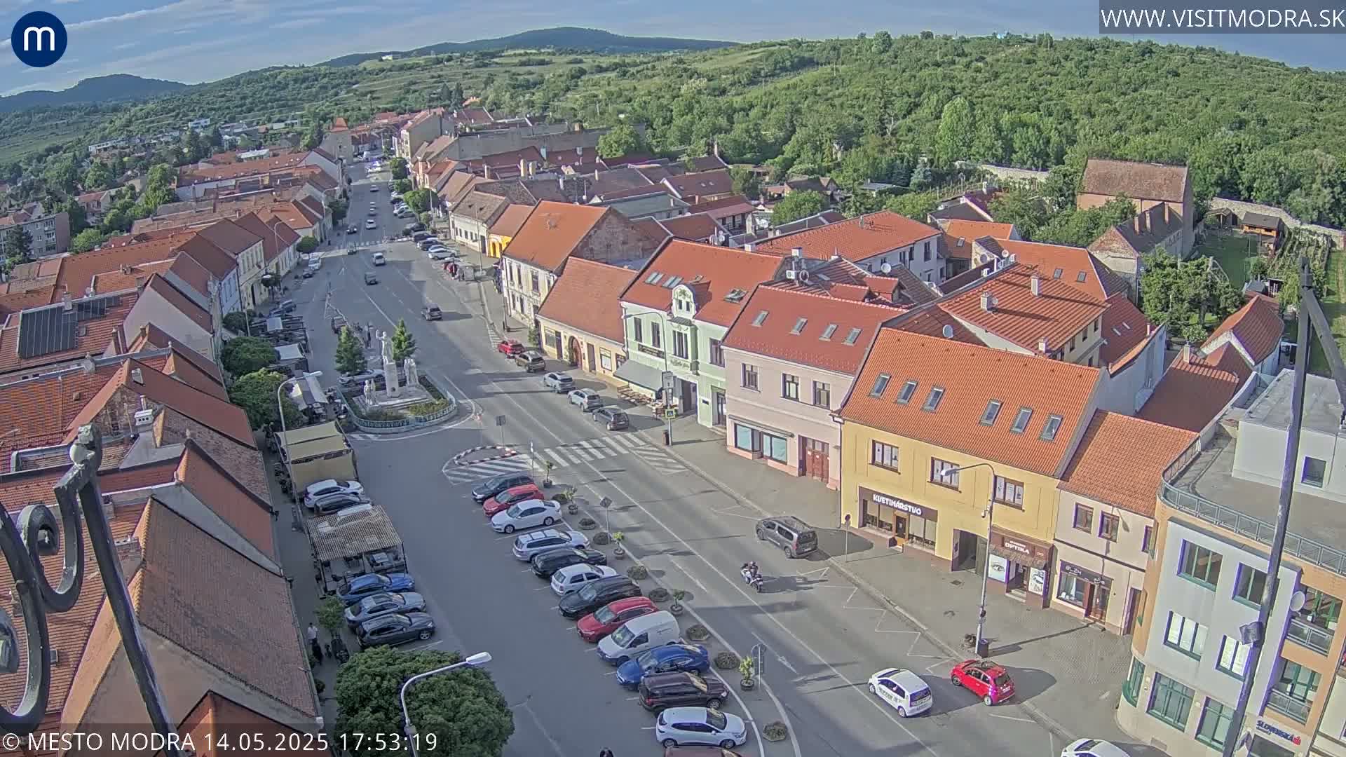 An aerial view of a town square on a sunny day, showing rows of parked cars lining a street flanked by colorful buildings.