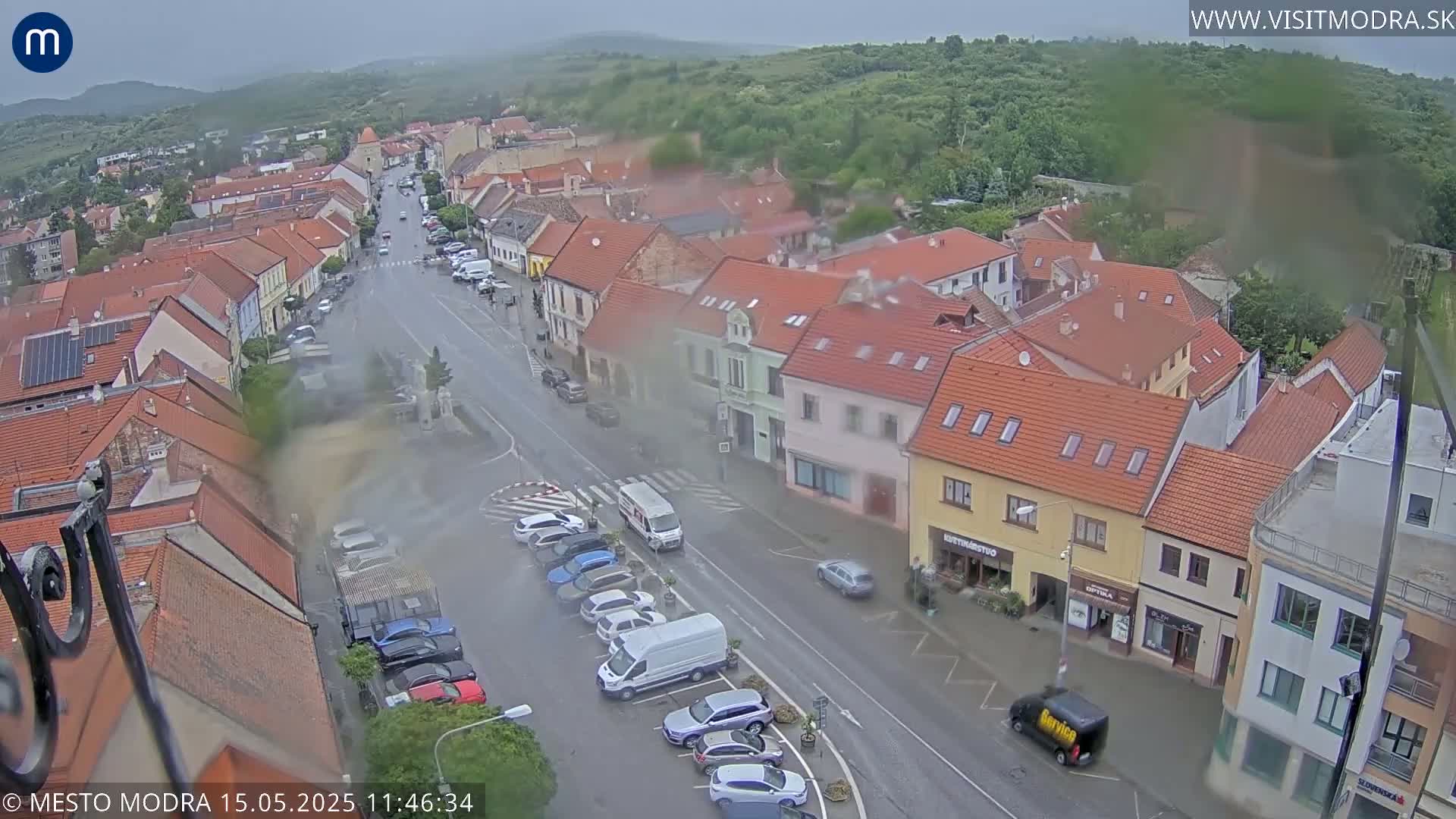 A rain-misted aerial view shows a town square with parked cars and several low buildings in a European style, surrounded by lush greenery on a cloudy day.