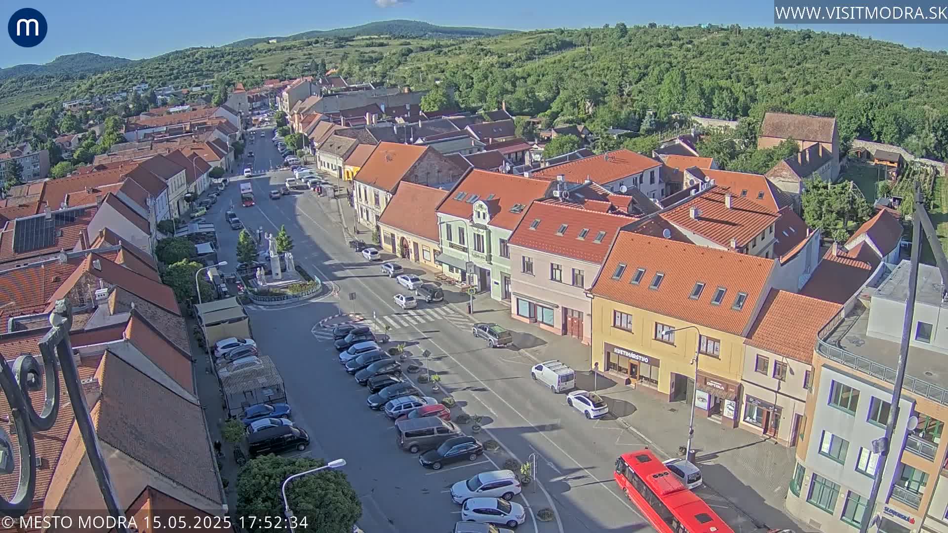 A sunny aerial view of a town square with parked cars, a bus, and buildings with red-tiled roofs.