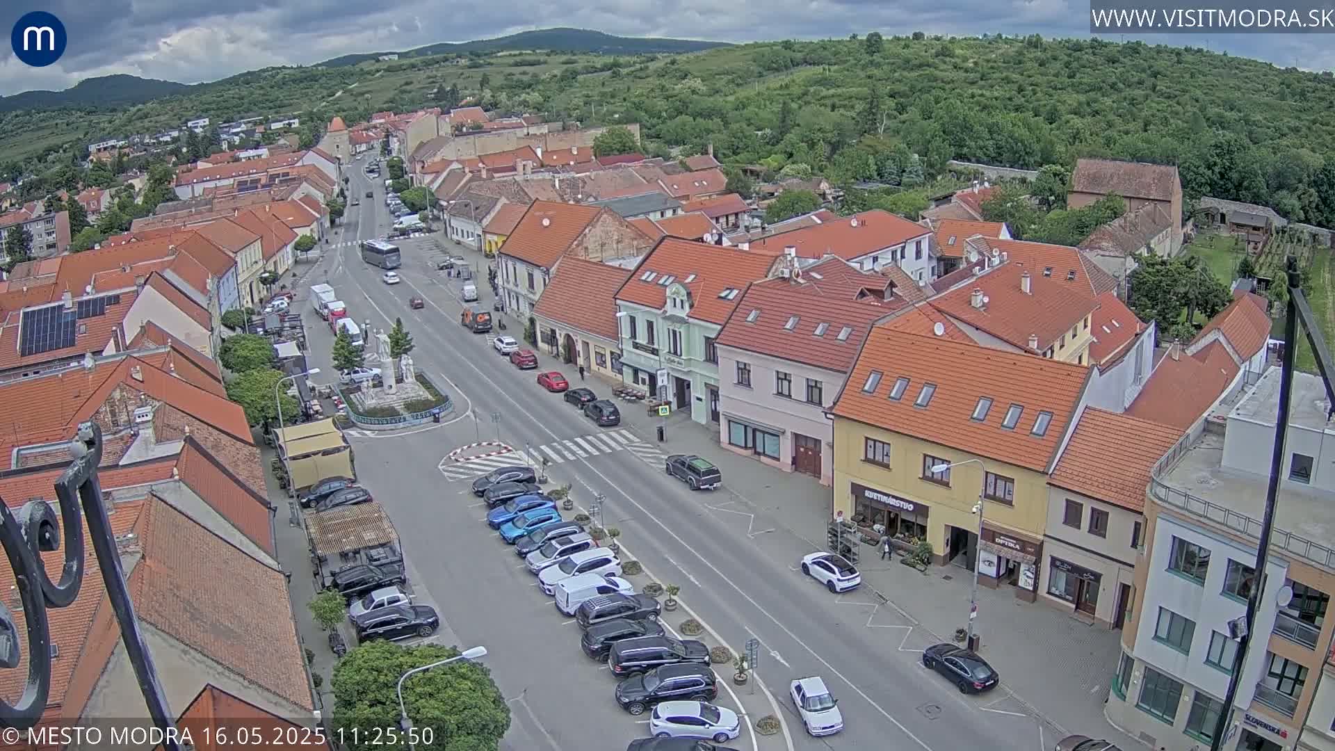 A town square with overcast skies and parked cars lines a street flanked by two-story buildings.