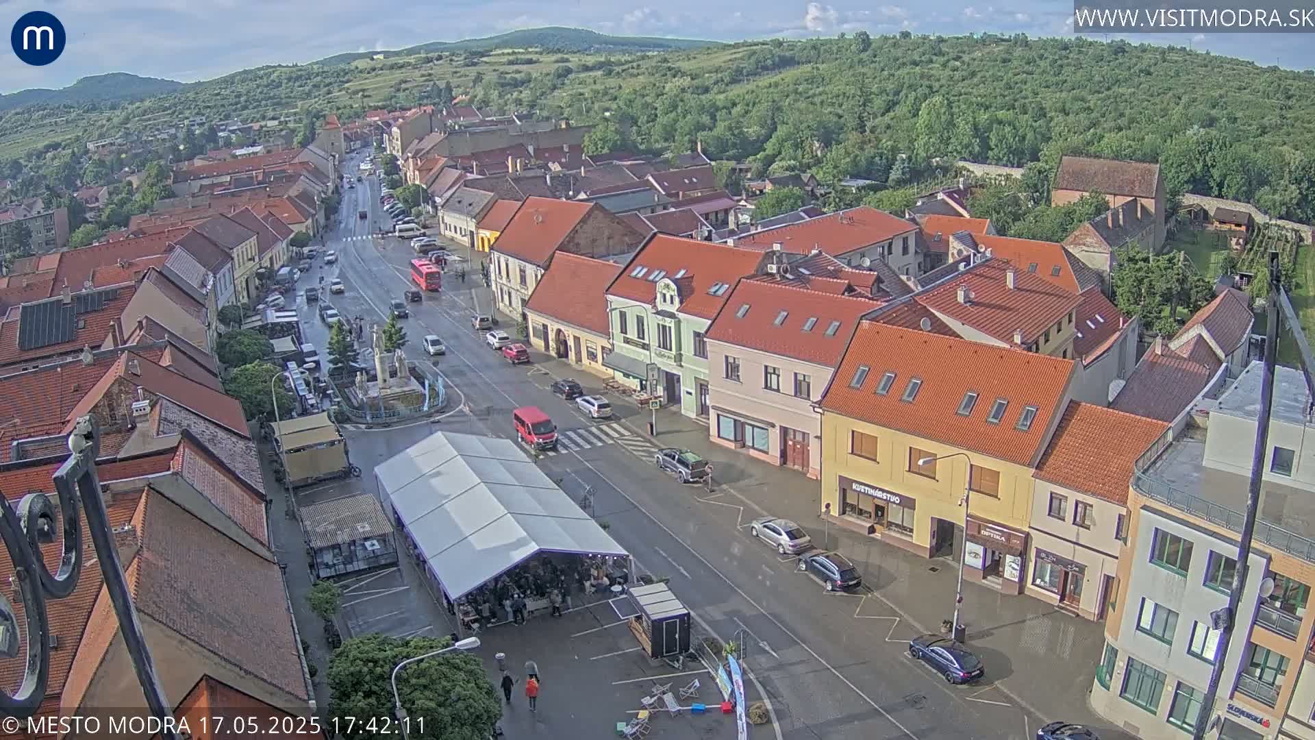 A high-angle view of a town square with red-roofed buildings, a street with cars, a market tent, and slightly wet roads under a partly cloudy sky.