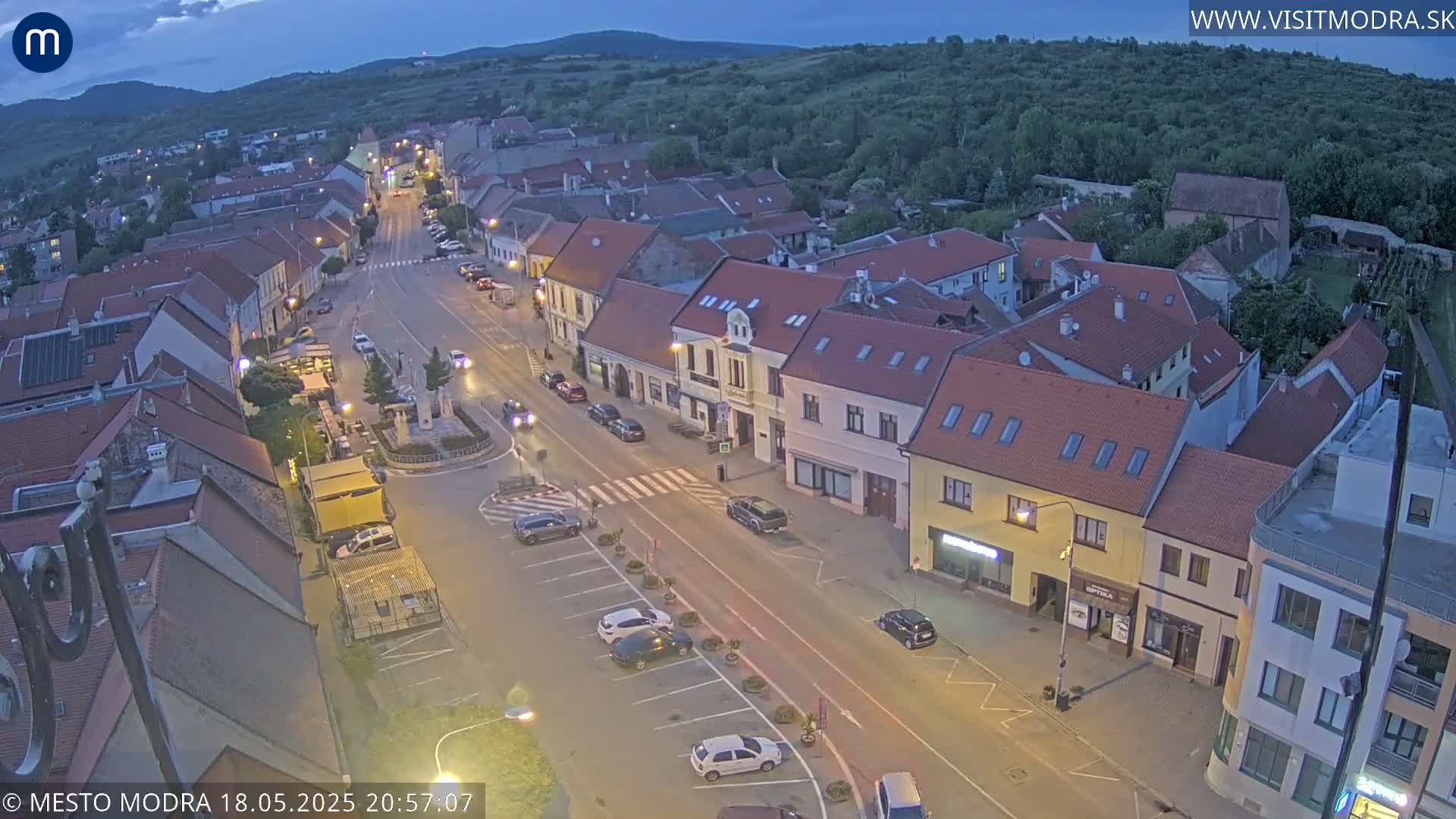 An aerial view at twilight shows a town square and surrounding streets with parked cars and buildings, under a mostly clear sky.