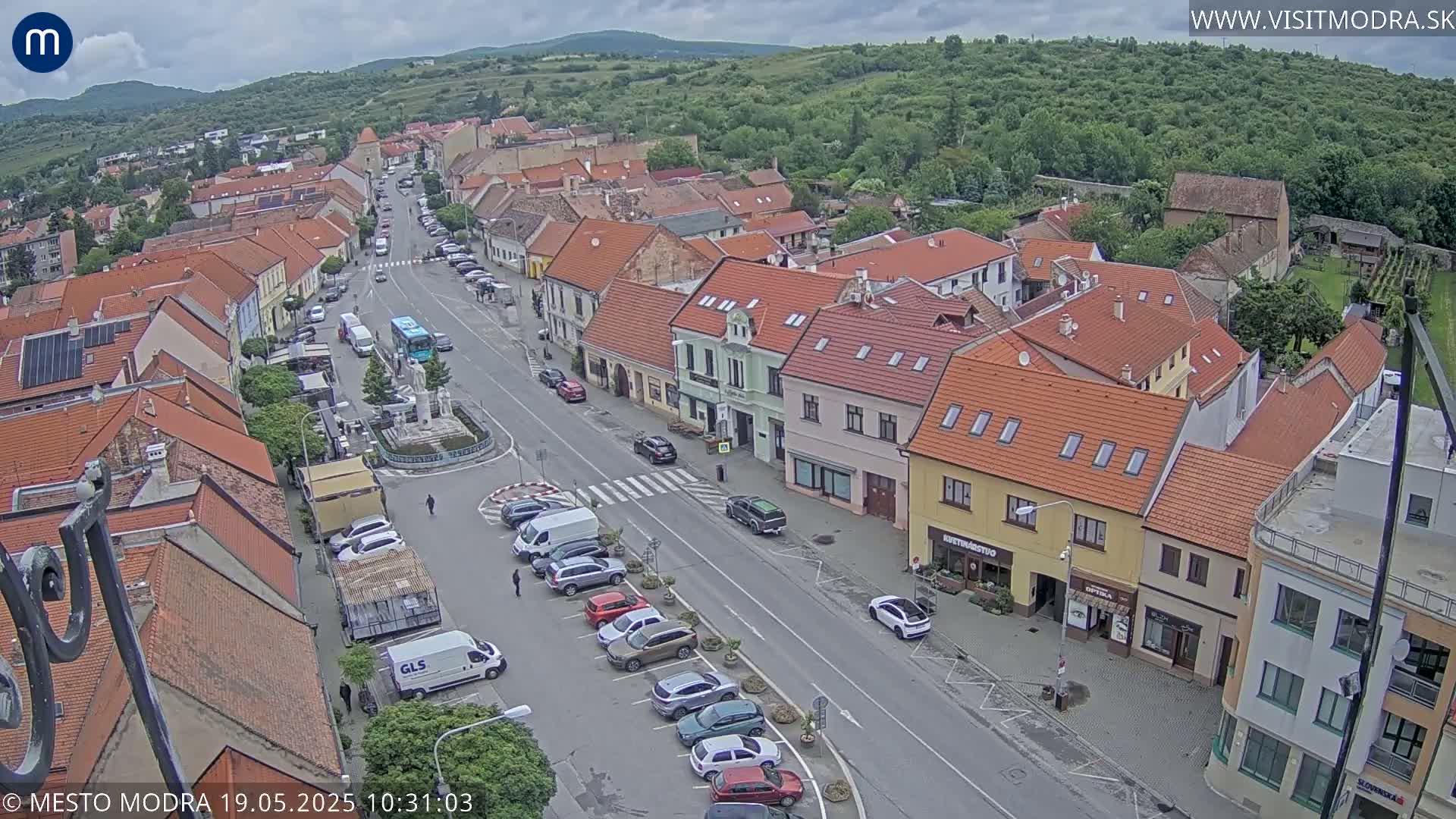 An overcast day reveals a bird's-eye view of a town square with red-roofed buildings, parked cars, and a bus, surrounded by lush green hills.