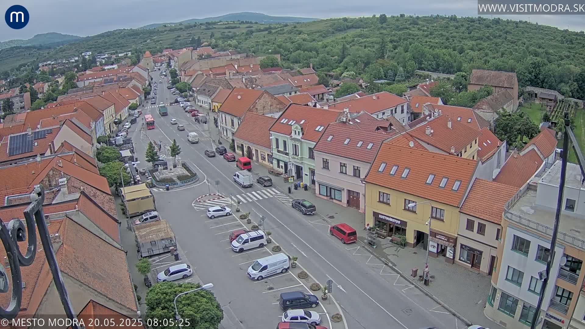 An aerial view of a town square and surrounding buildings on an overcast day, showing parked cars, a few moving vehicles, and a small town with red-tiled roofs.