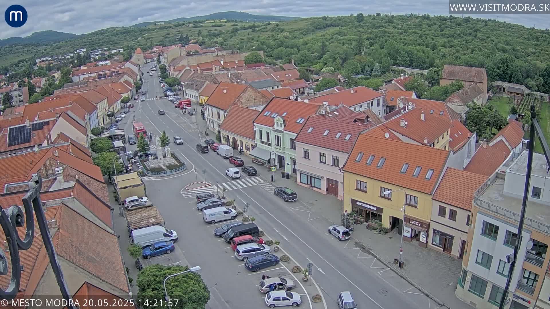 An aerial view of a town under a partly cloudy sky shows a main street with parked cars and buildings with orange tile roofs.