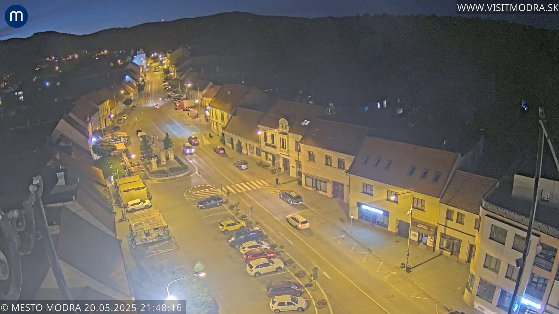 A nighttime aerial view of a town square with cars parked along the streets and a few vehicles driving, under a clear, dark sky.