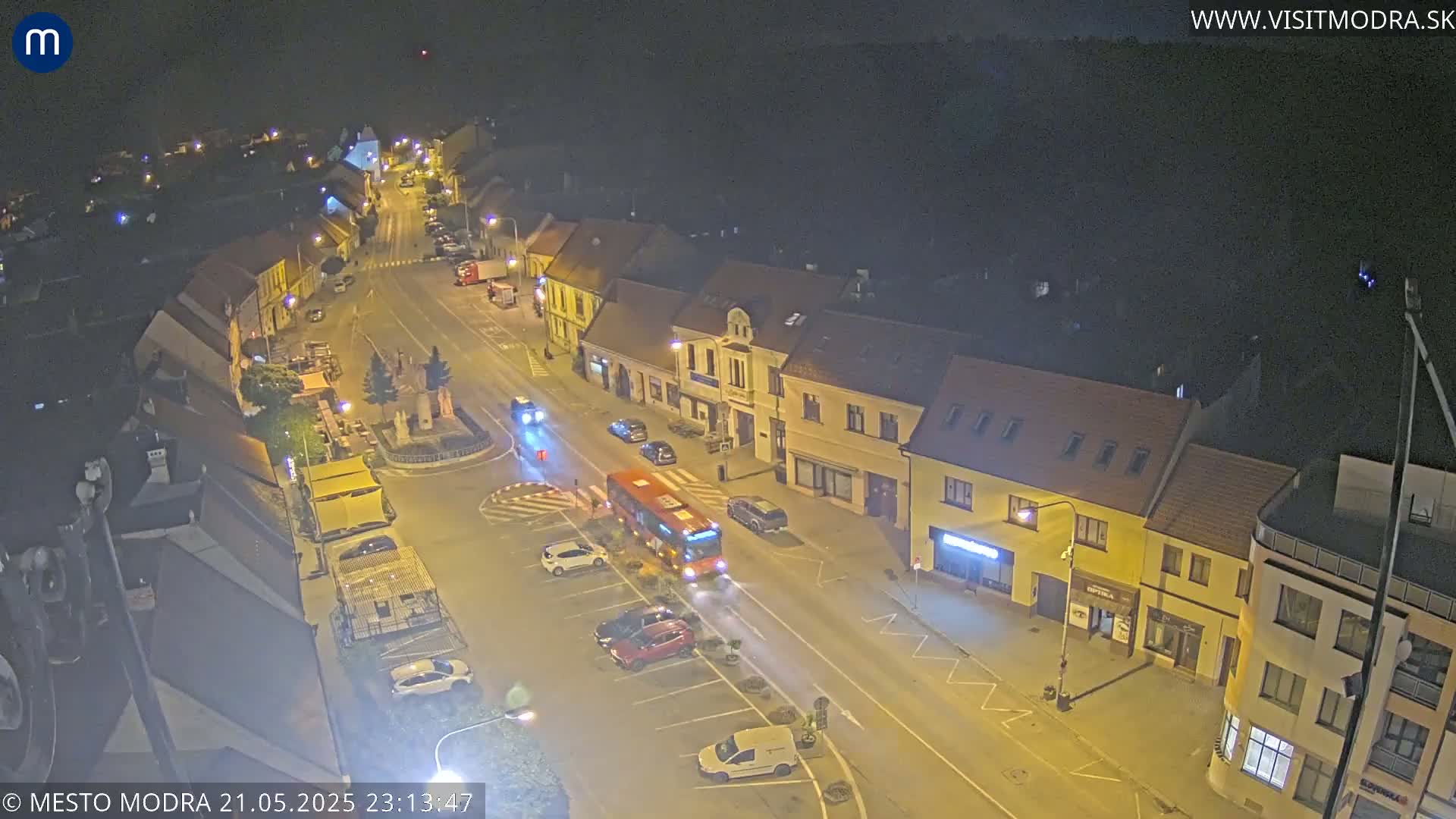 An elevated nighttime view of a town square with cars parked along the street and a bus traveling down the road, under a clear sky.