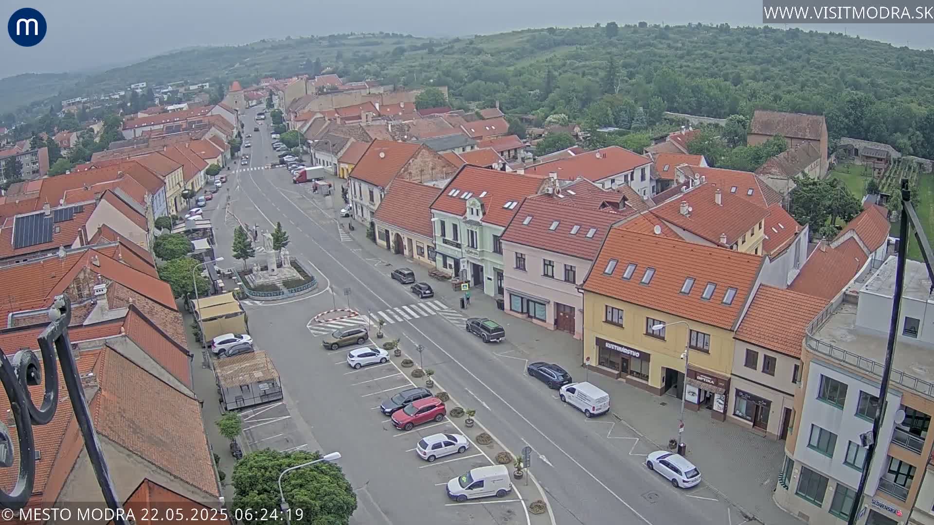 An overcast day reveals a bird's-eye view of a town's main street, lined with buildings featuring red-tiled roofs and parked cars.