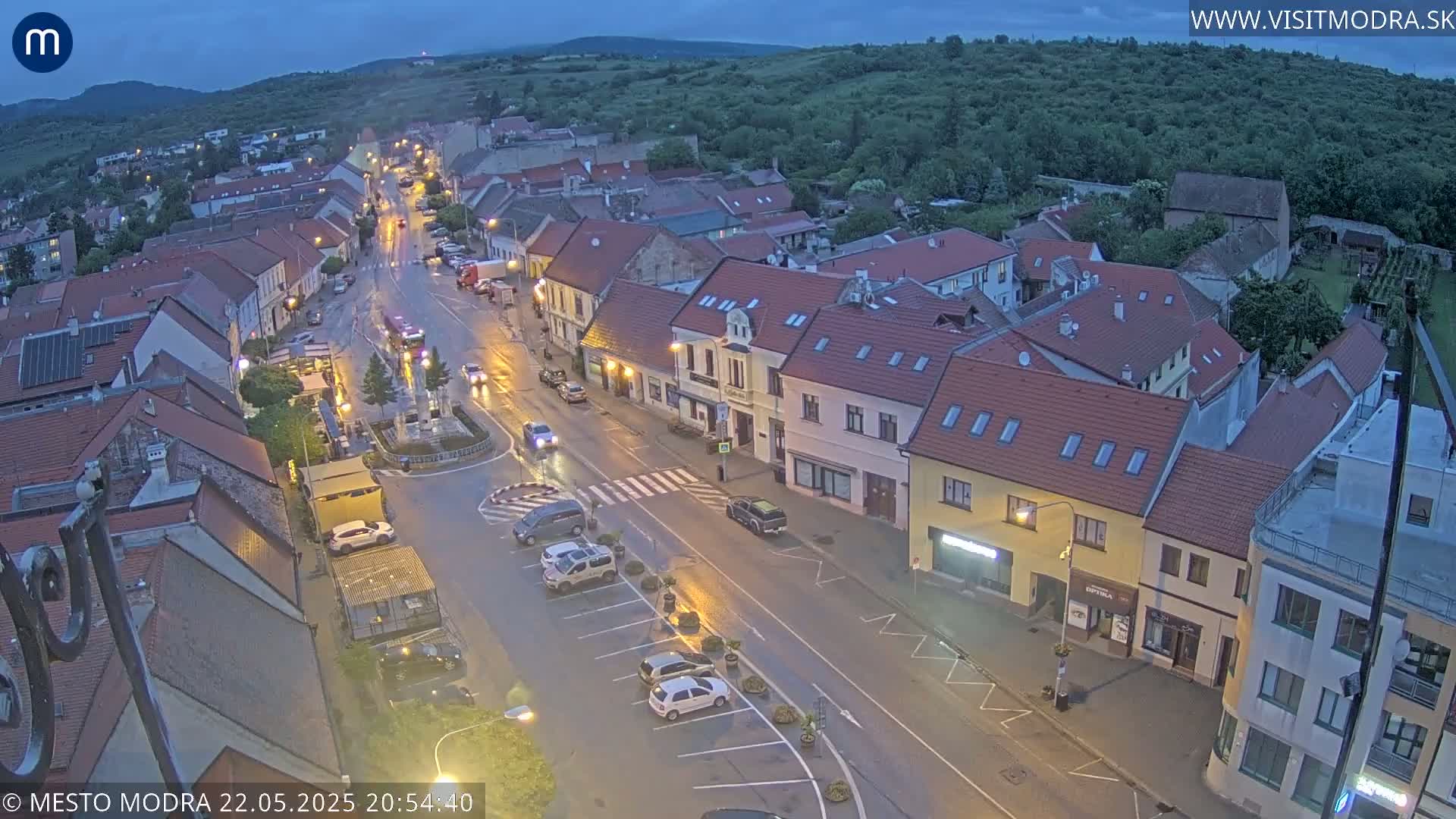 An aerial view of a town at twilight, showing a main street with parked cars and buildings lining both sides, under a mostly clear sky.