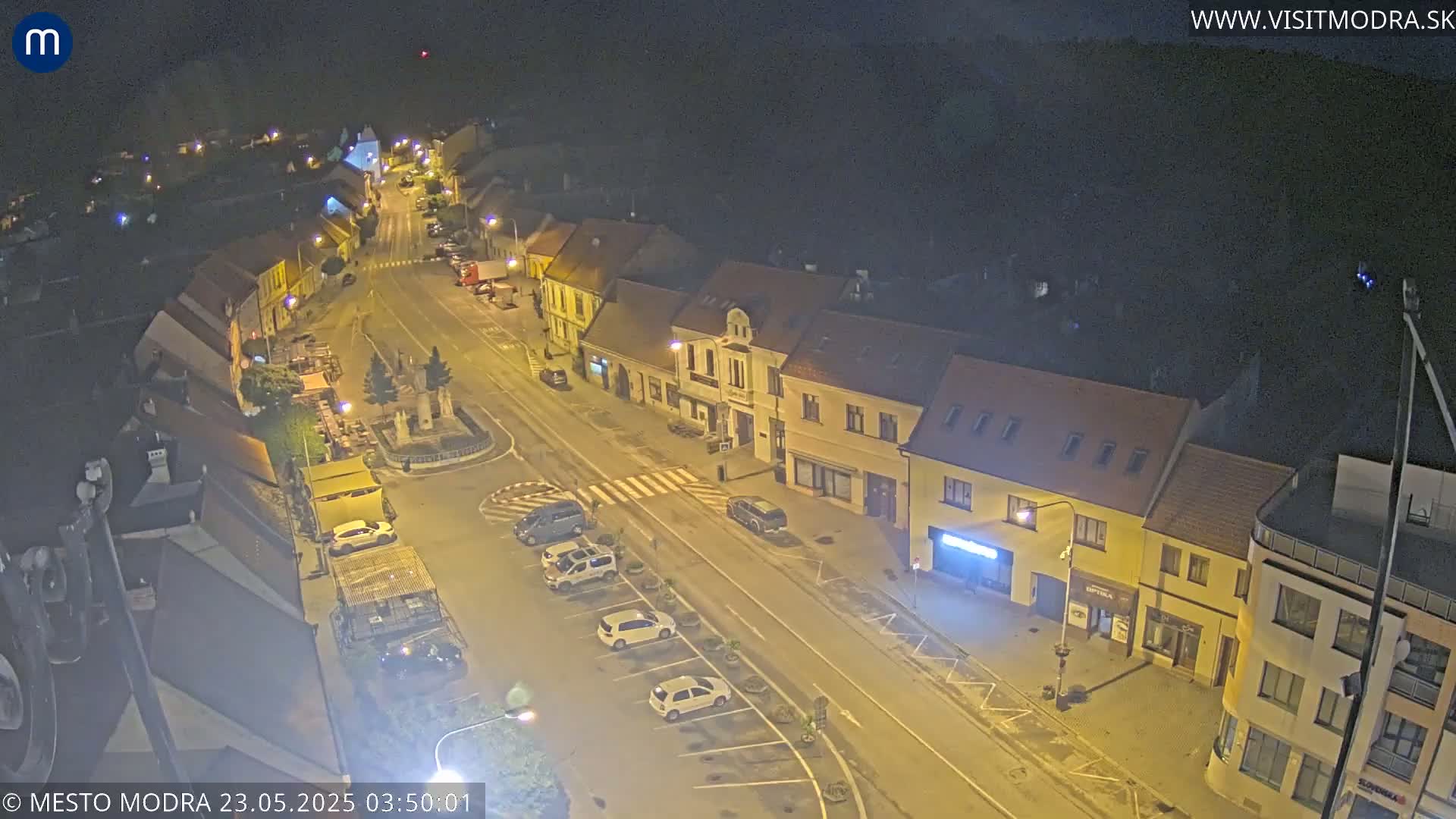A nighttime, high-angle view of a town square with parked cars and buildings illuminated by streetlights.