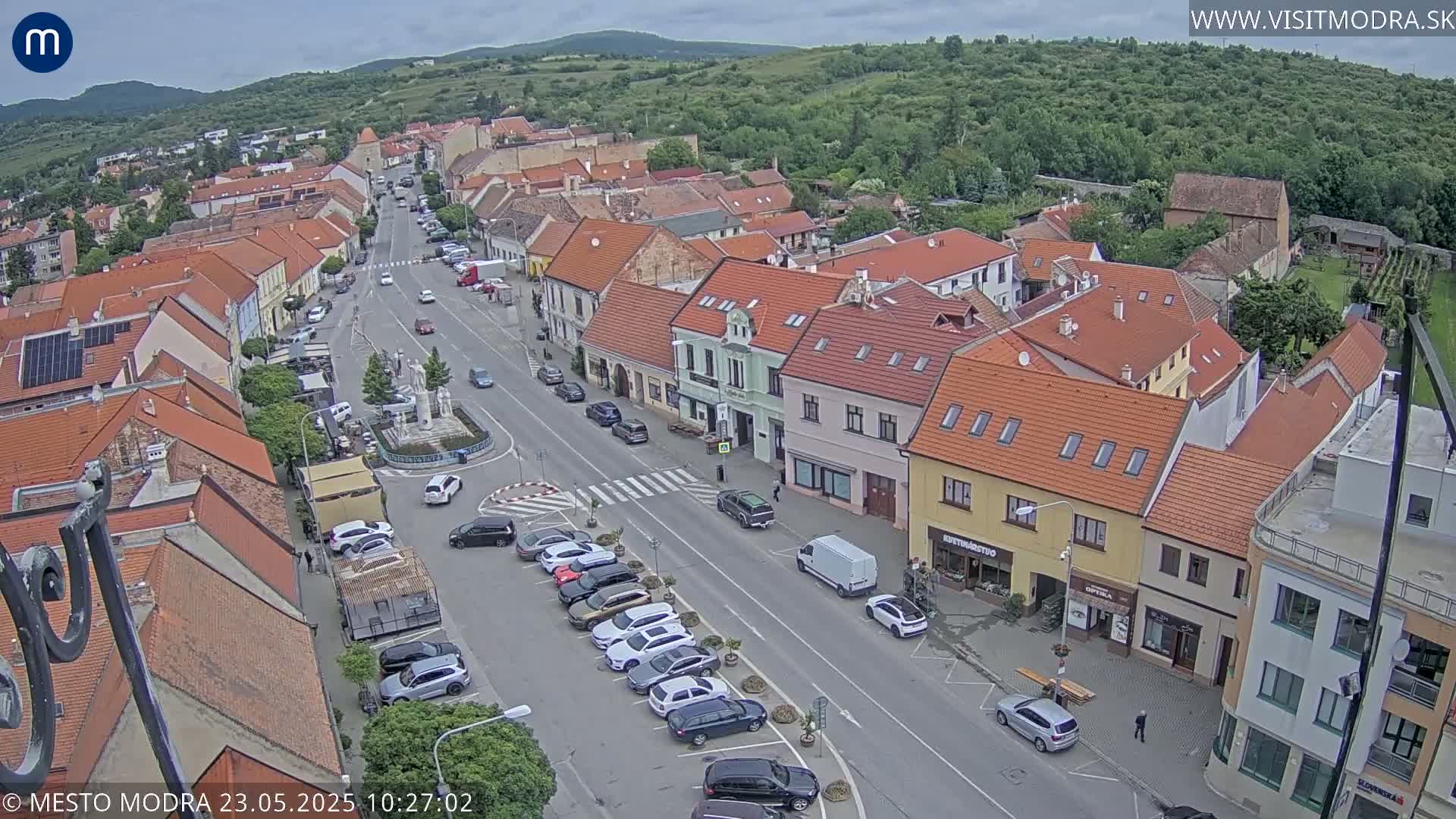 An aerial view of a town's main street on a partly cloudy day, showing parked cars, buildings with red tile roofs, and a small central square.