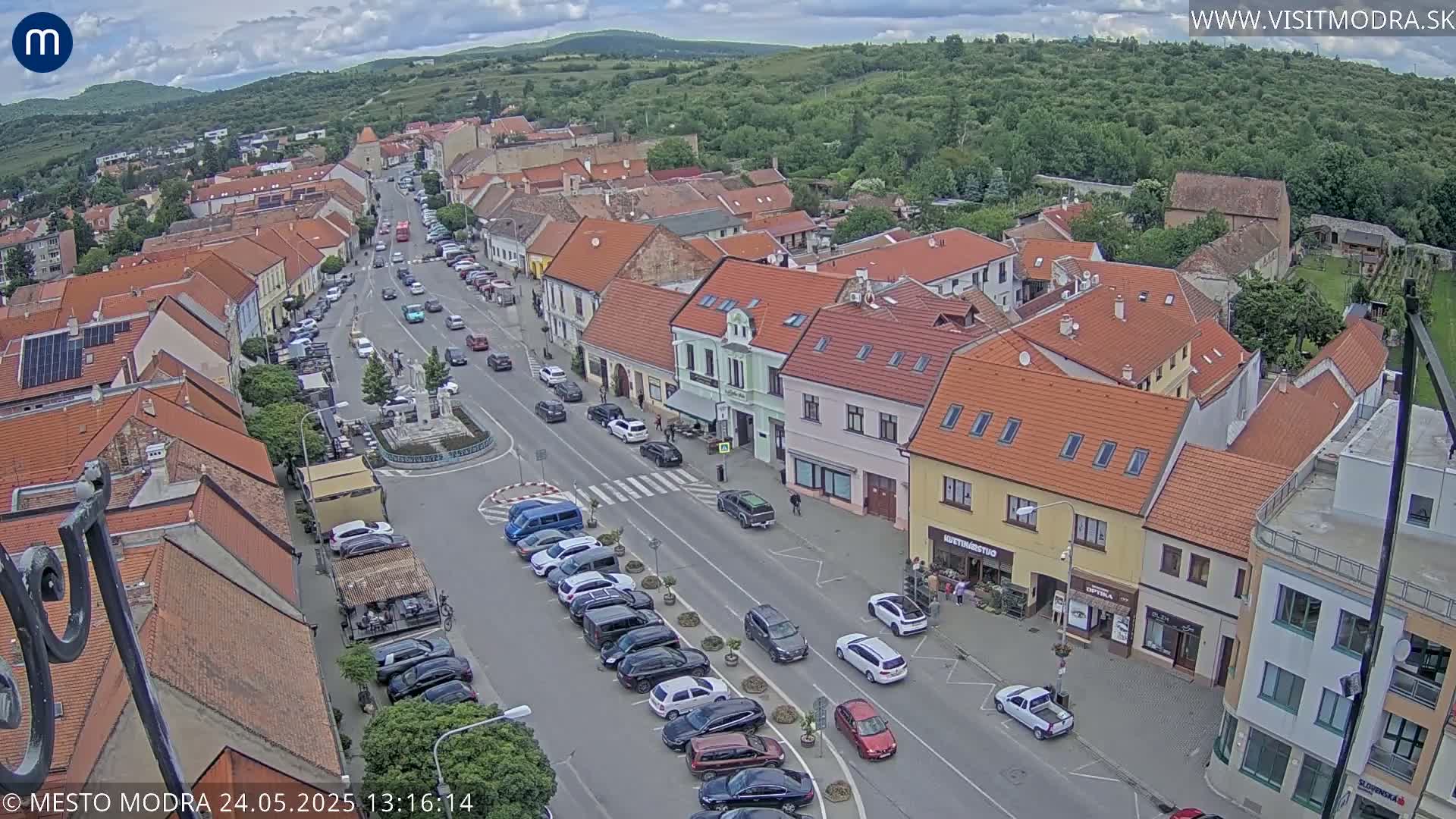 An aerial view of a town's main street on a partly cloudy day, showing parked cars lining the street and other vehicles driving on the road, with buildings of various sizes and styles surrounding a central roundabout.