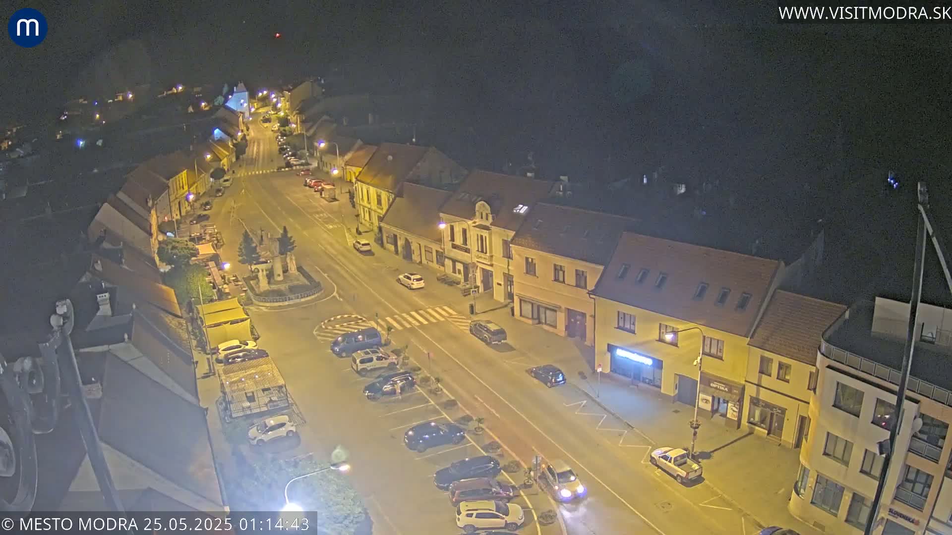 A nighttime, high-angle view shows a town square with parked cars and buildings lining a street under clear skies.