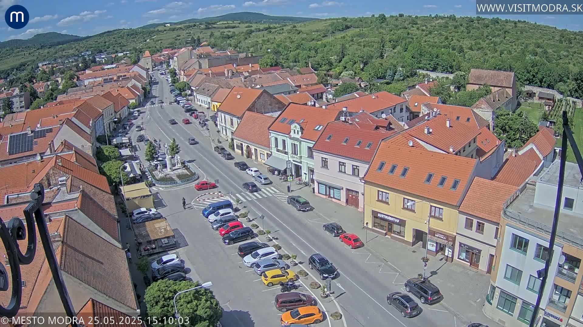 An aerial view of a town square on a sunny day shows a street lined with parked cars and buildings with red-tiled roofs.
