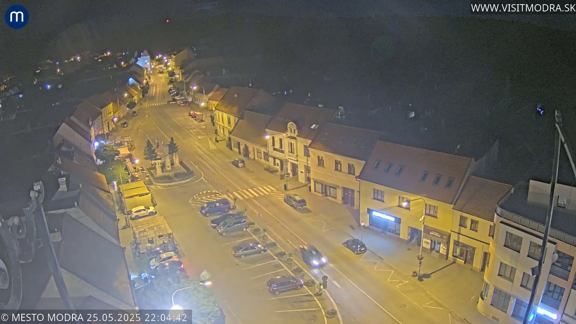 A nighttime, high-angle view of a town square with several cars parked along the streets and buildings lit by streetlights.