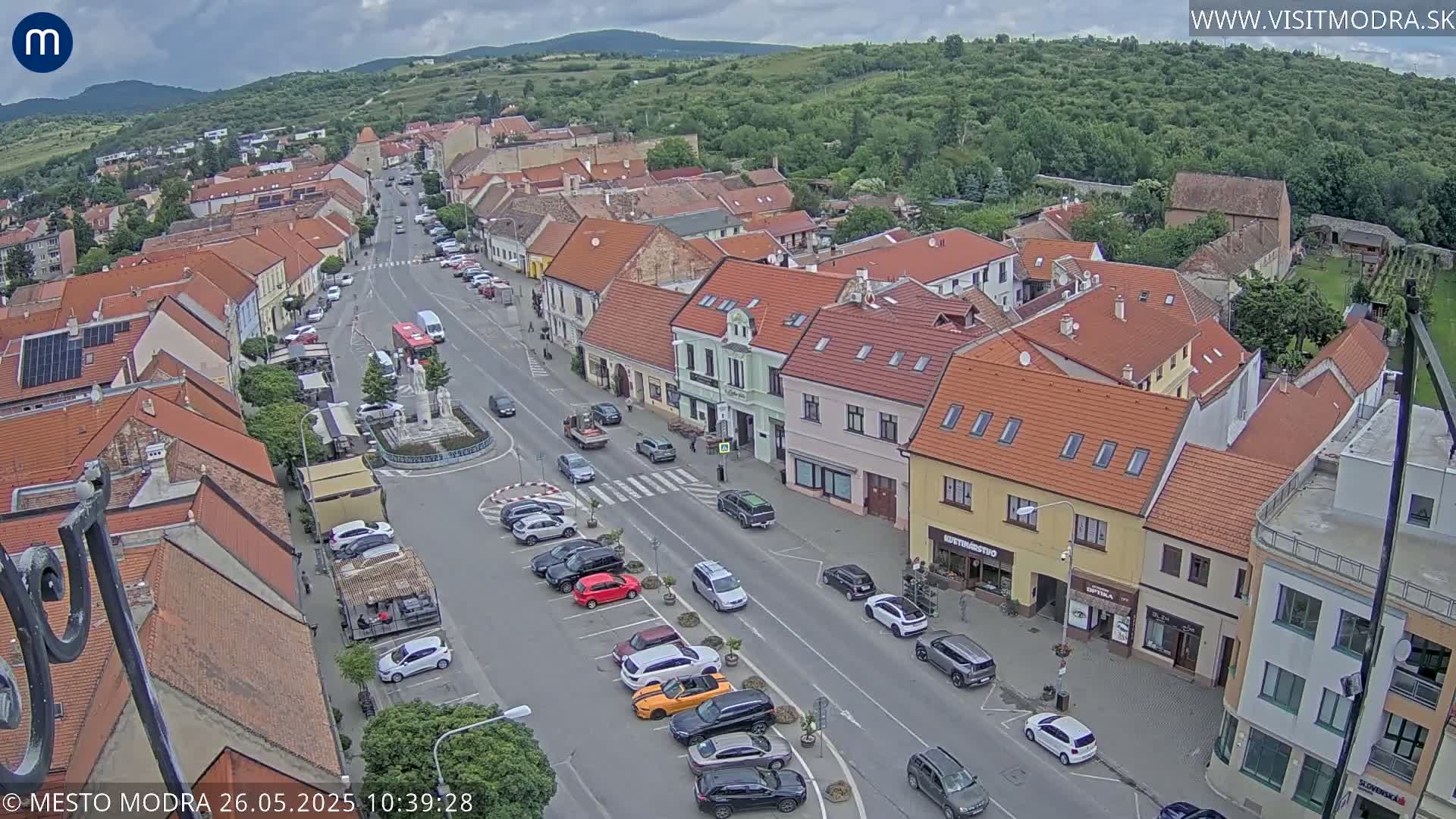 An aerial view of a town square on a partly cloudy day, showing parked cars along a street lined with buildings and a central roundabout with a fountain.