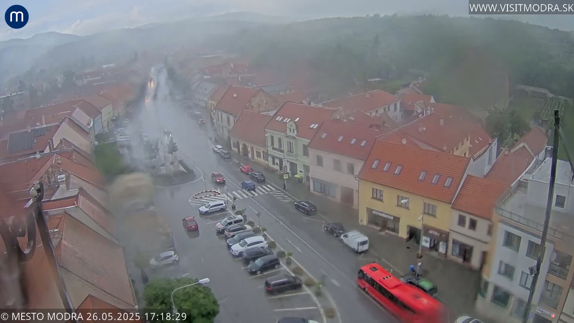 A high-angle view of a town square with cars and a bus on a rain-swept street, amidst buildings with red tile roofs, under a hazy sky.