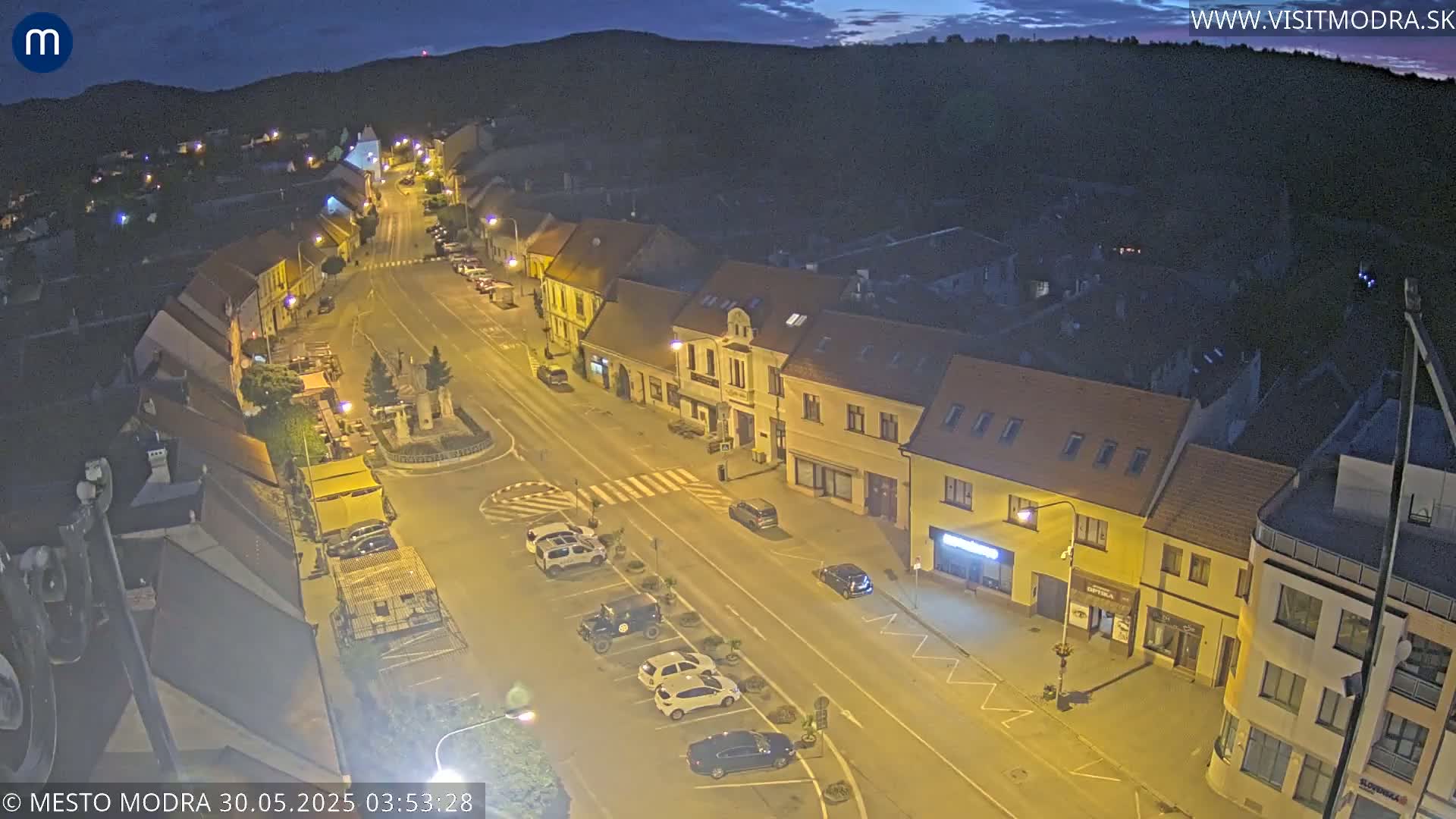 A nighttime, high-angle view of a town square with cars parked along the street and buildings lit by streetlights.