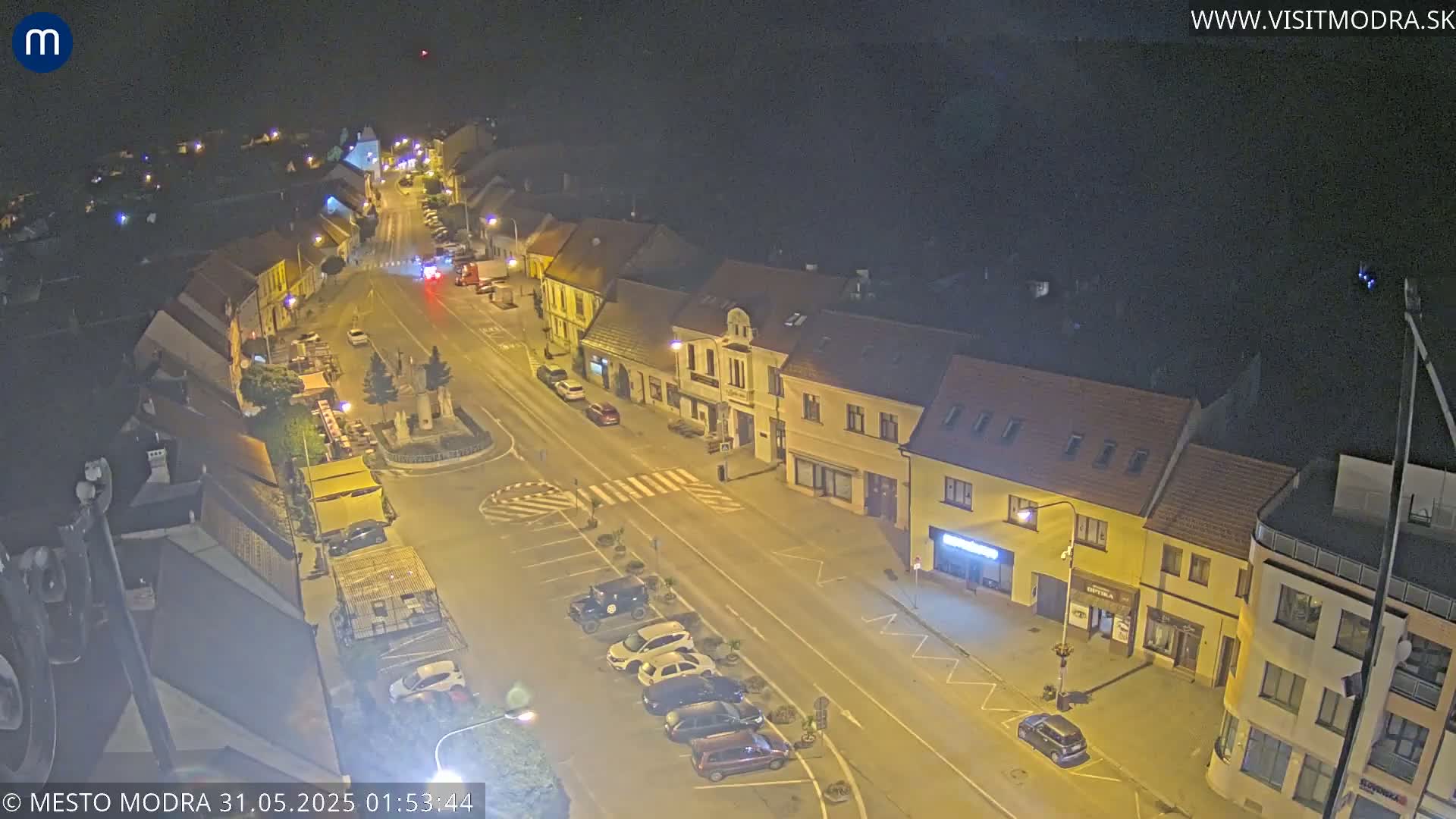 A nighttime, high-angle view of a town square with several cars parked along the street and buildings lining the road under a clear, dark sky.
