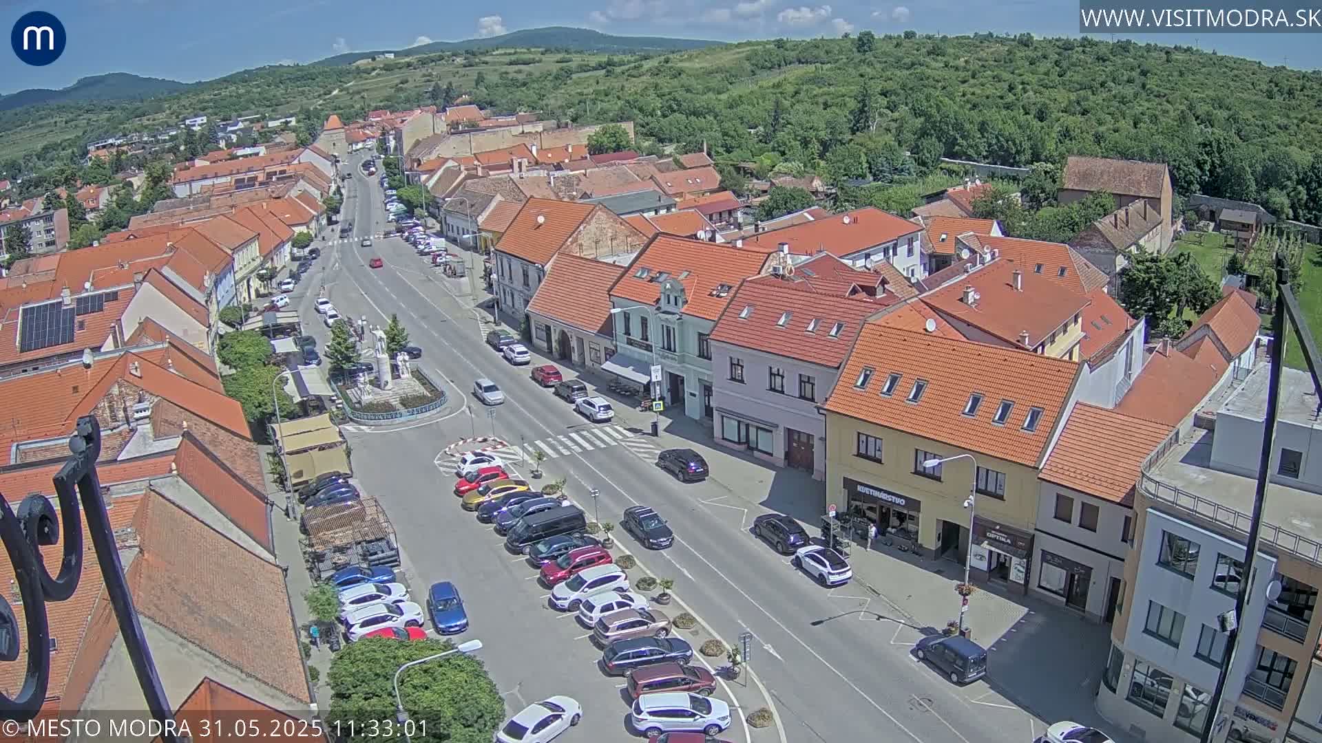 A sunny aerial view of a town square and surrounding streets lined with buildings, many with red tile roofs, and parked cars.
