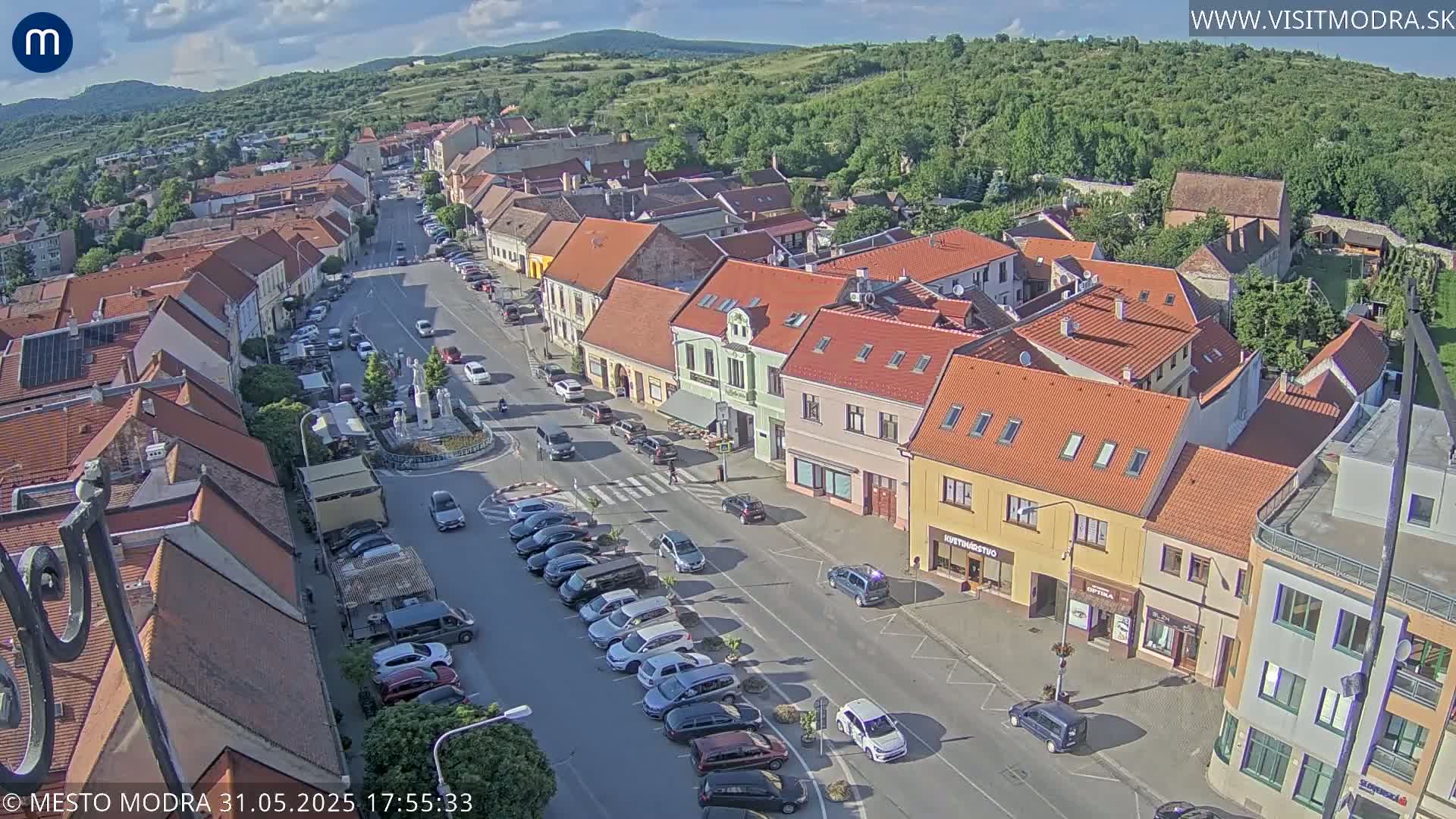 A mostly sunny aerial view of a town square with parked cars and buildings lining a main street, surrounded by greenery and hills in the background.