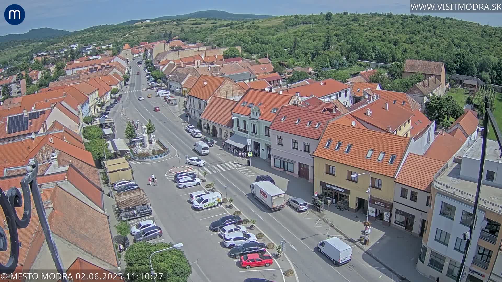 A sunny day overlooks a town square with parked cars and buildings with red tile roofs.
