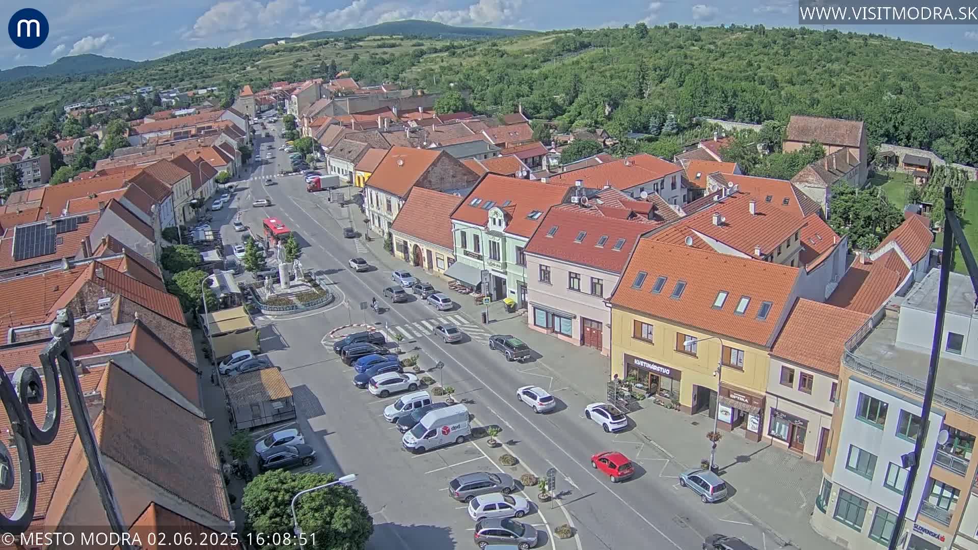 A town square with red-roofed buildings and parked cars is seen on a partly sunny day, with hills visible in the background.