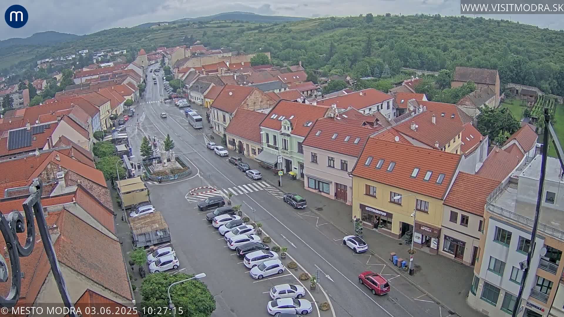 An aerial view of a town square with overcast skies, showing a main street lined with parked cars and buildings with red-tiled roofs.