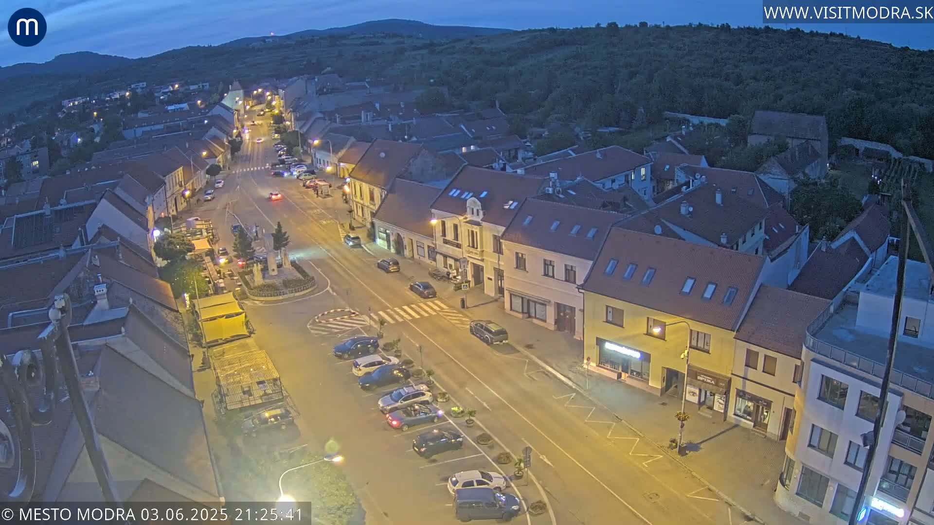 An aerial view at dusk shows a town's street lined with buildings, cars parked along the sides, and a few vehicles on the road.