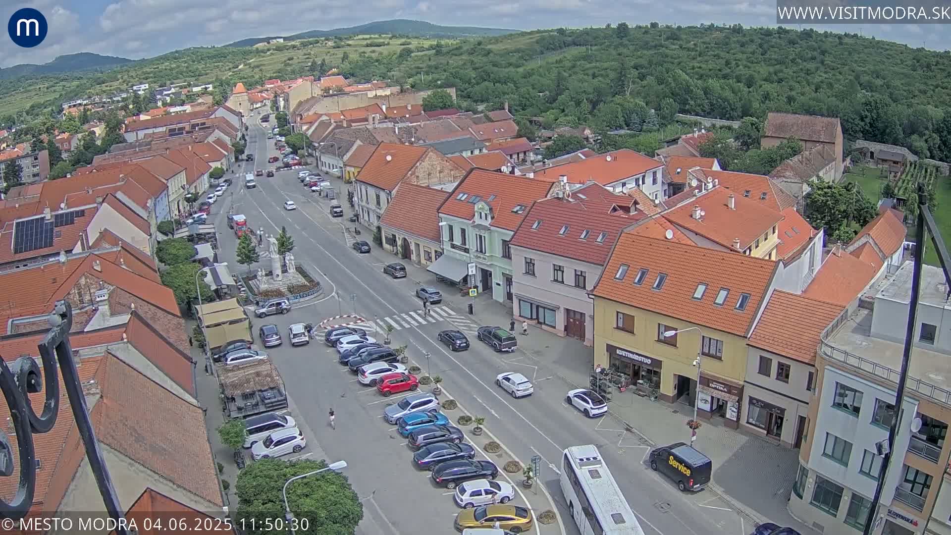 A partly cloudy day reveals a high-angle view of a town square with parked cars lining a main road,  surrounded by buildings with red tile roofs, and hills in the background.