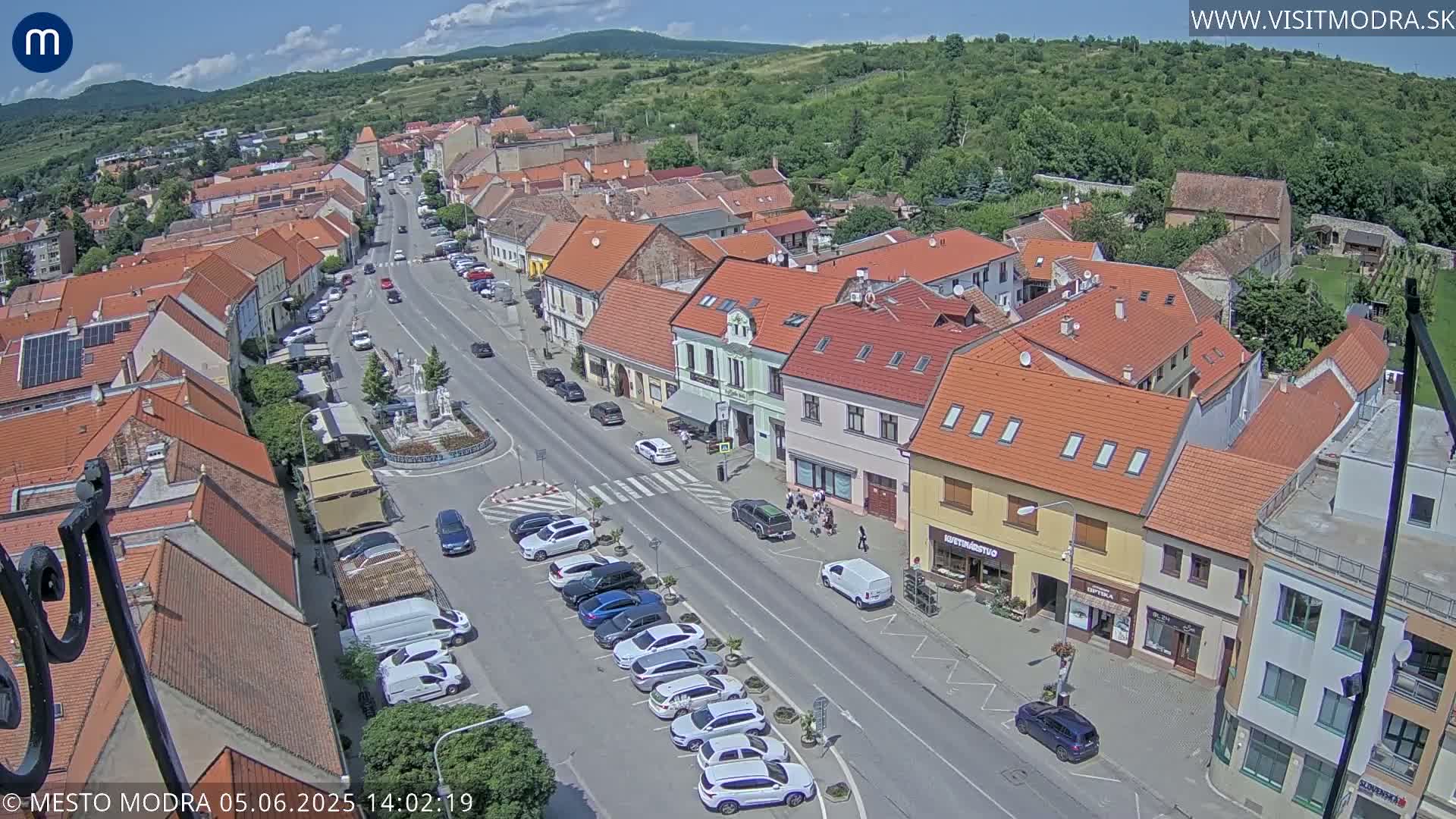 A high-angle, sunny-day view of a town's main street lined with parked cars and low-rise buildings, some with red-tiled roofs.