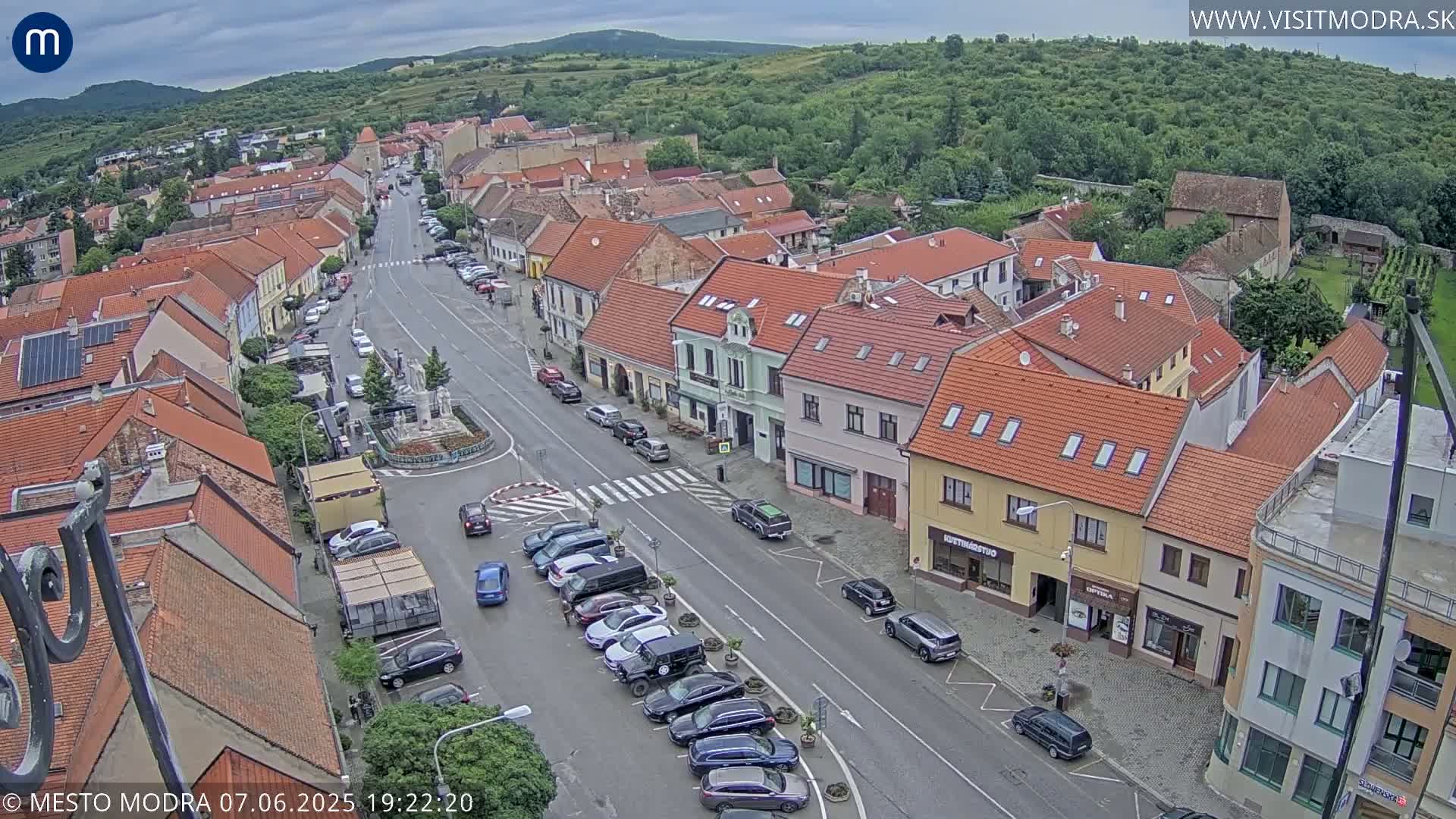 An aerial view of a town square with overcast skies, showing a main street lined with parked cars and buildings with red-tiled roofs.