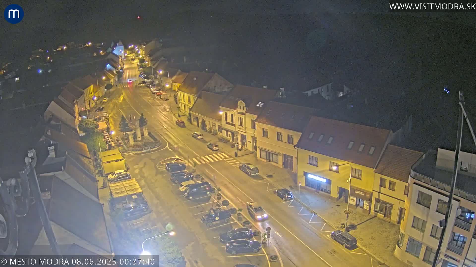 A nighttime, high-angle view of a town square with parked cars lining the streets, illuminated by streetlights.