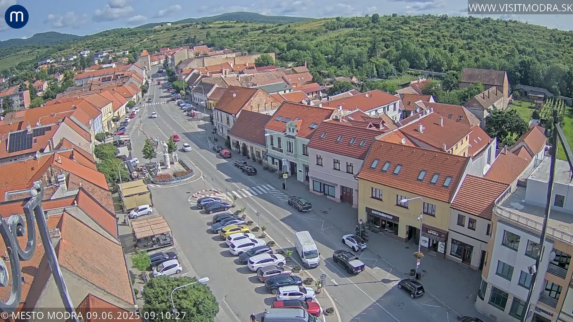 A sunny aerial view of a town square with parked cars lining a street, surrounded by buildings with red tile roofs.