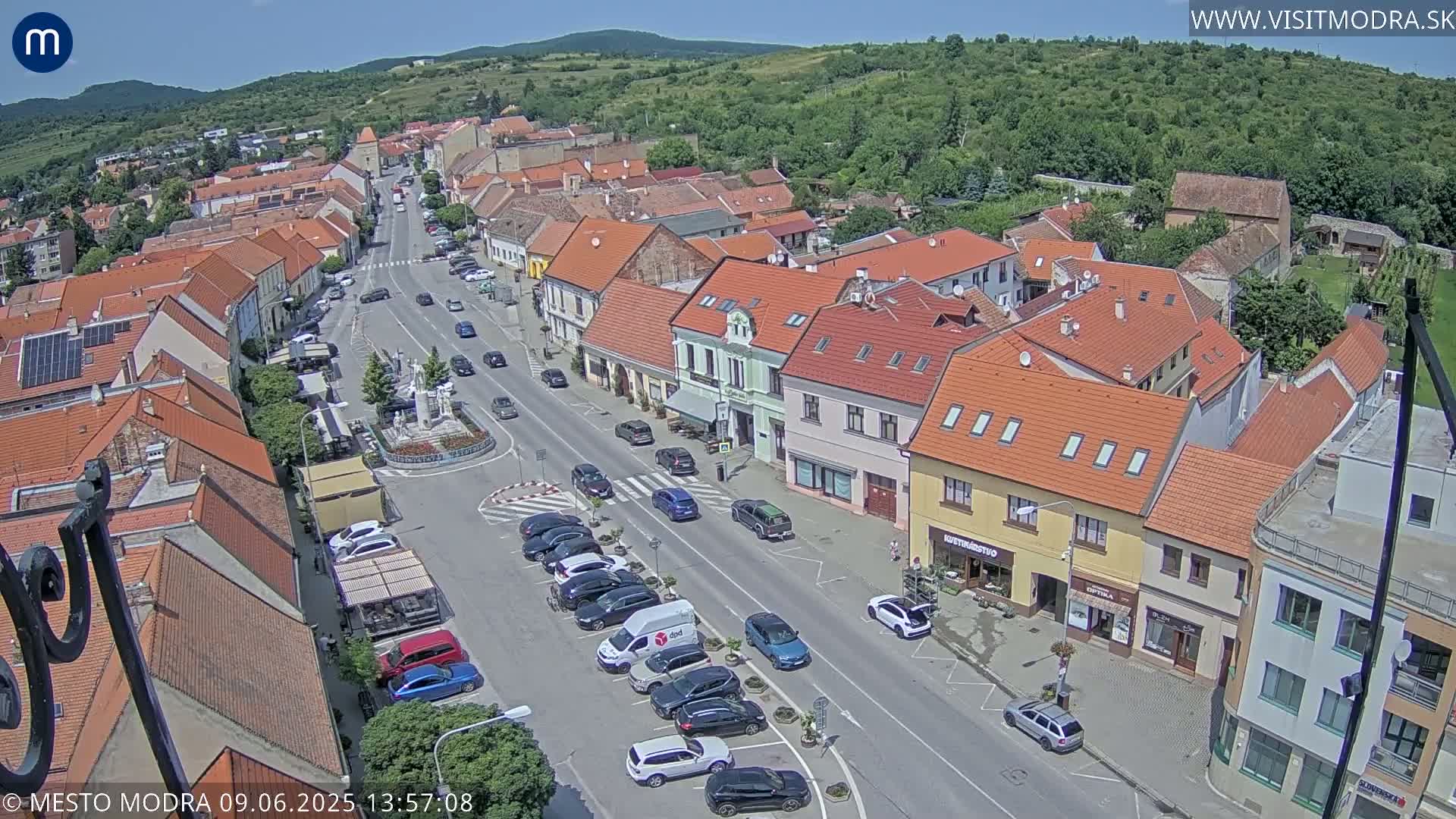 An aerial view of a town square and surrounding buildings on a sunny day, showing parked cars and a few people walking.