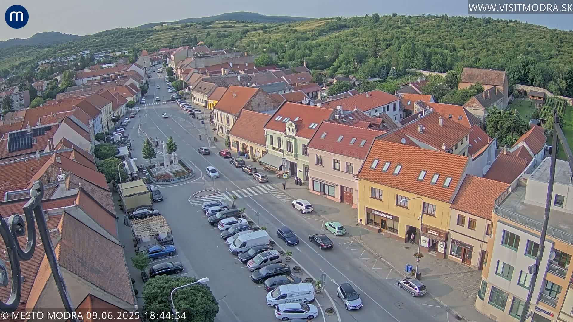 An aerial view of a town square with parked cars, buildings with red tile roofs, and a central statue under a clear evening sky.