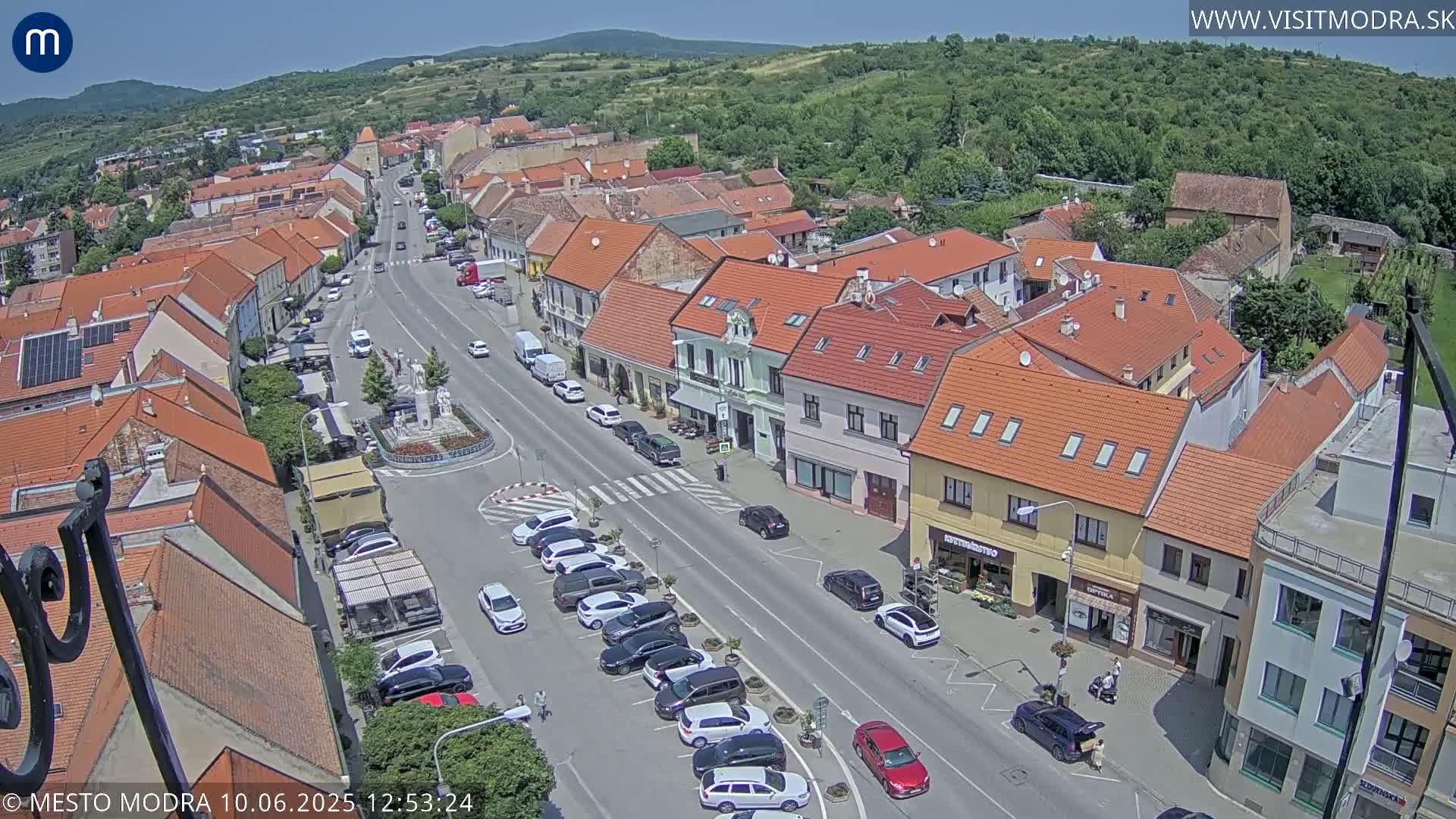 An aerial view of a town square on a sunny day, showing buildings with red tile roofs and cars parked along a main street.