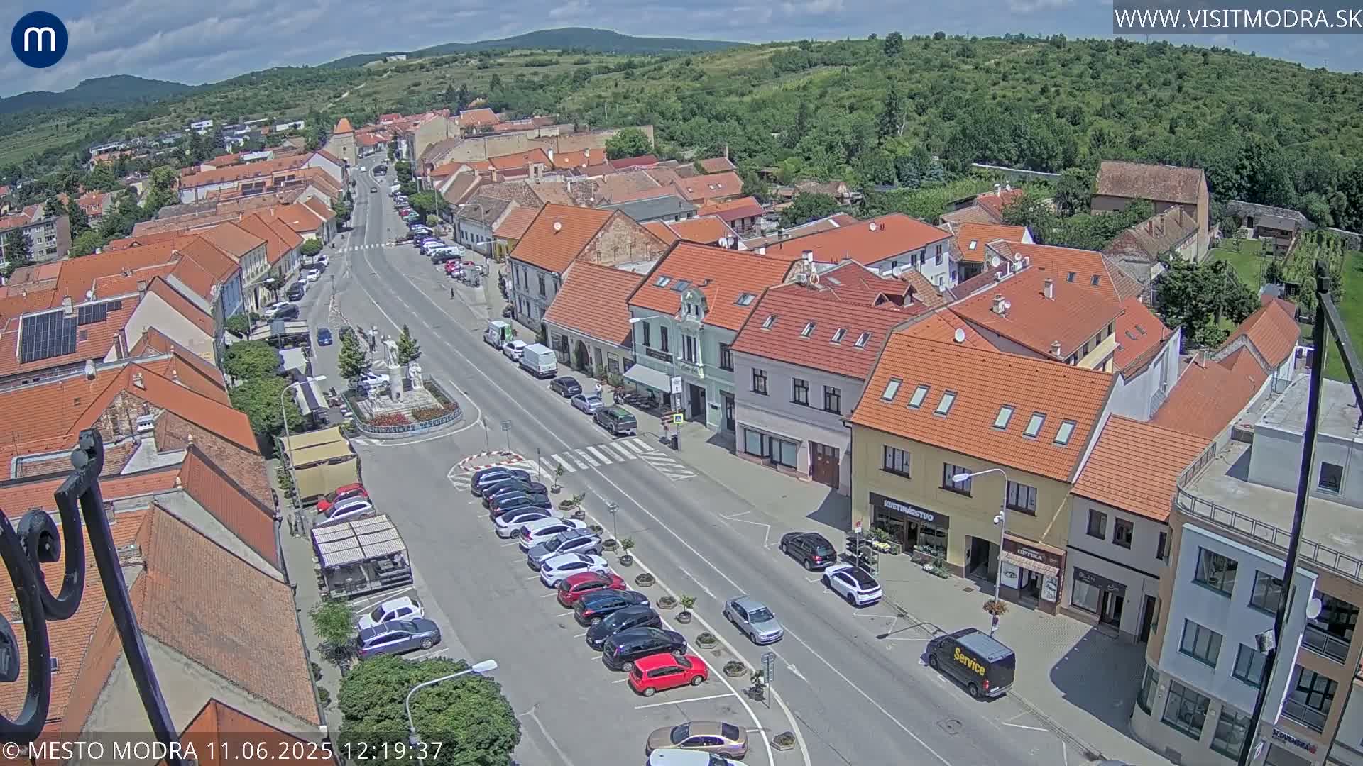 A high-angle, sunny view of a town square and street lined with parked cars and buildings with red tile roofs, nestled at the foot of a verdant hill.
