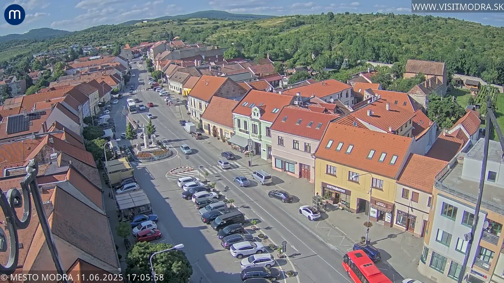 An aerial view of a town square on a sunny day, showing a street lined with buildings and parked cars, along with a small roundabout in the center.