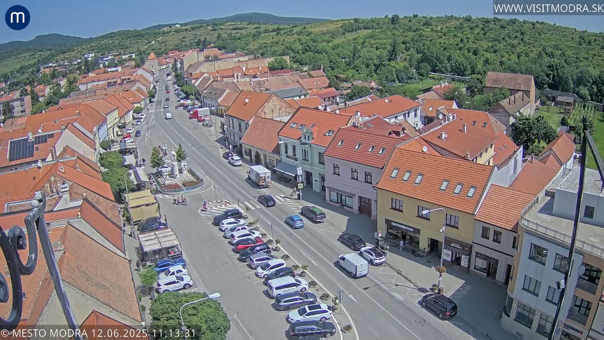 An aerial view of a town on a sunny day shows a main street lined with parked cars and buildings, with a small town square visible in the middle.
