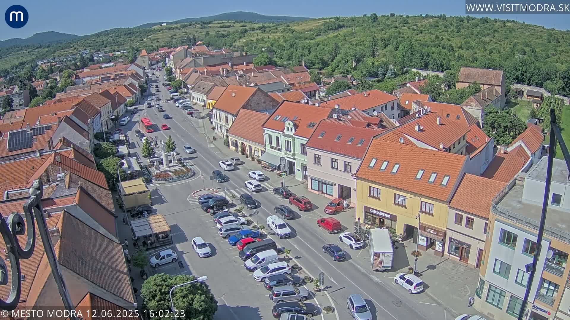 A sunny aerial view of a town's main street, lined with parked cars and buildings, leading towards a central square with a fountain.