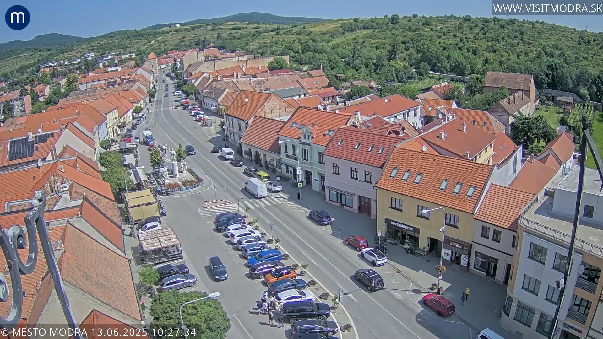 A sunny day reveals a town's main street lined with parked cars and traditional buildings, nestled against a verdant hillside.