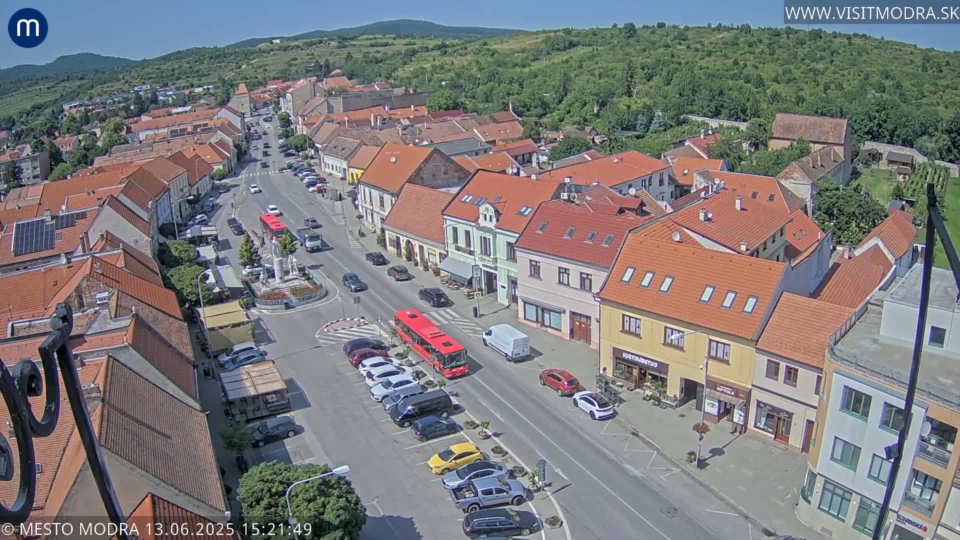An aerial view of a town's main street on a sunny day, showing parked cars, a red bus, and buildings with red tile roofs.