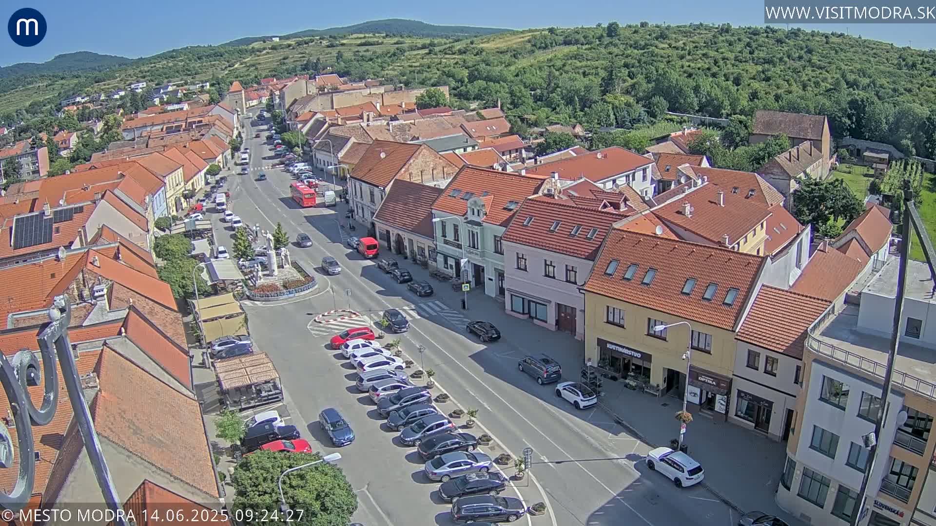 A high-angle, sunny view of a town's main street lined with parked cars and low-rise buildings, under a clear sky.