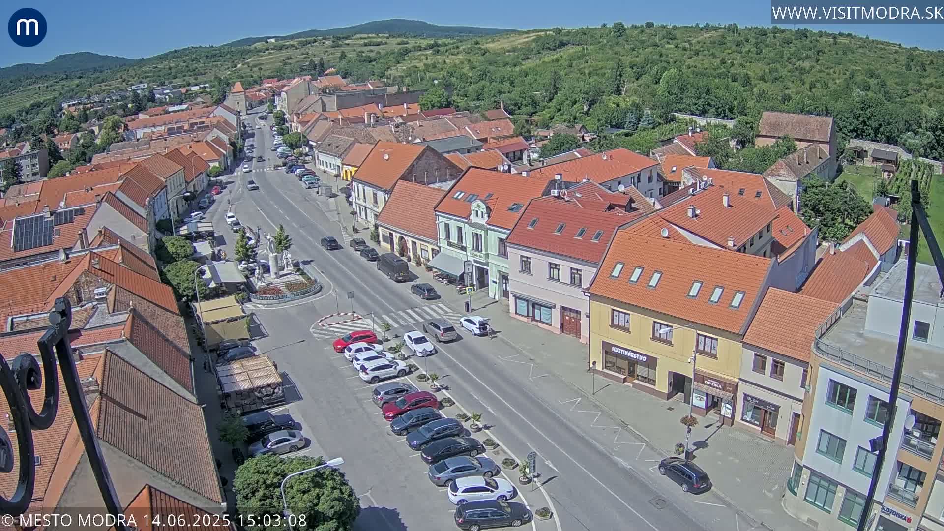 An aerial view of a town on a sunny day shows a main street lined with buildings, parked cars, and a small town square with a statue.