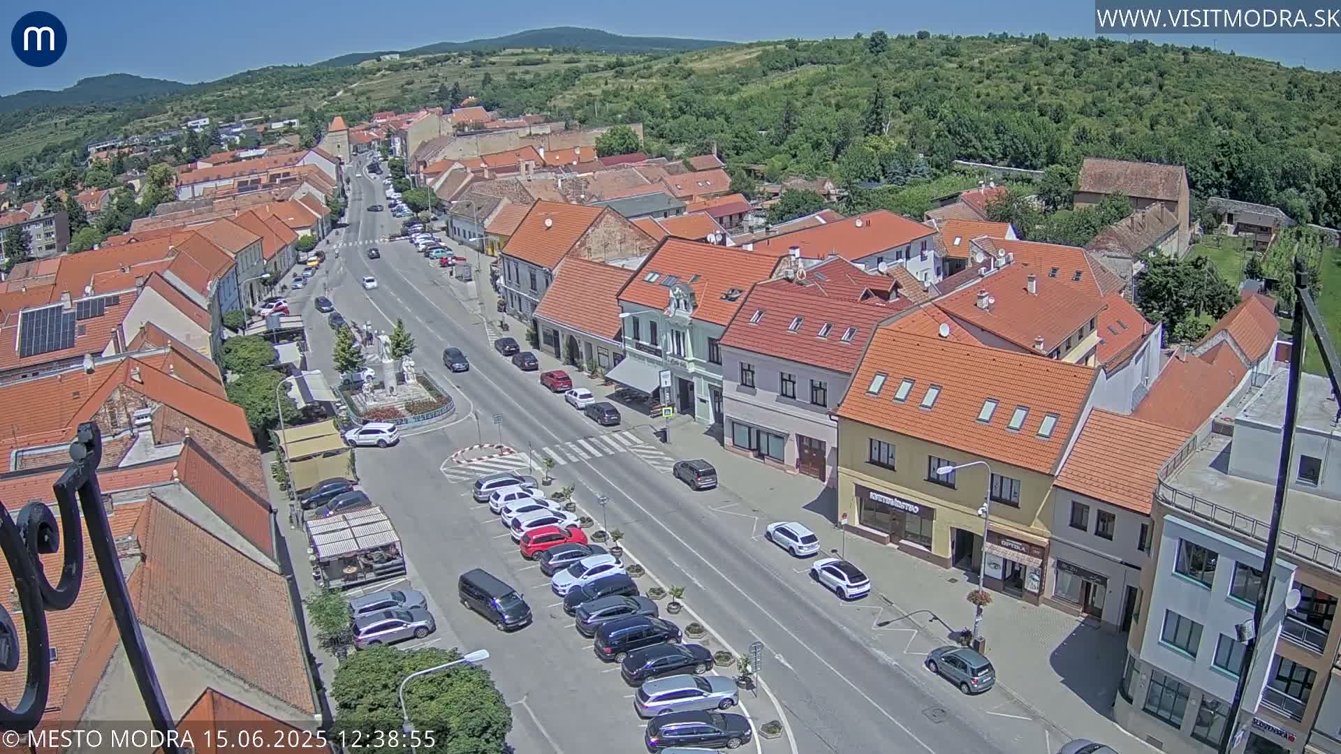 An aerial view of a town square on a sunny day, showing red-roofed buildings lining a street with parked cars.