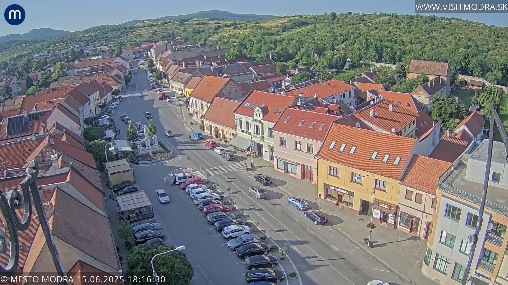 An aerial view shows a town square with parked cars lining a street and buildings with red-tiled roofs under a clear, sunny sky.
