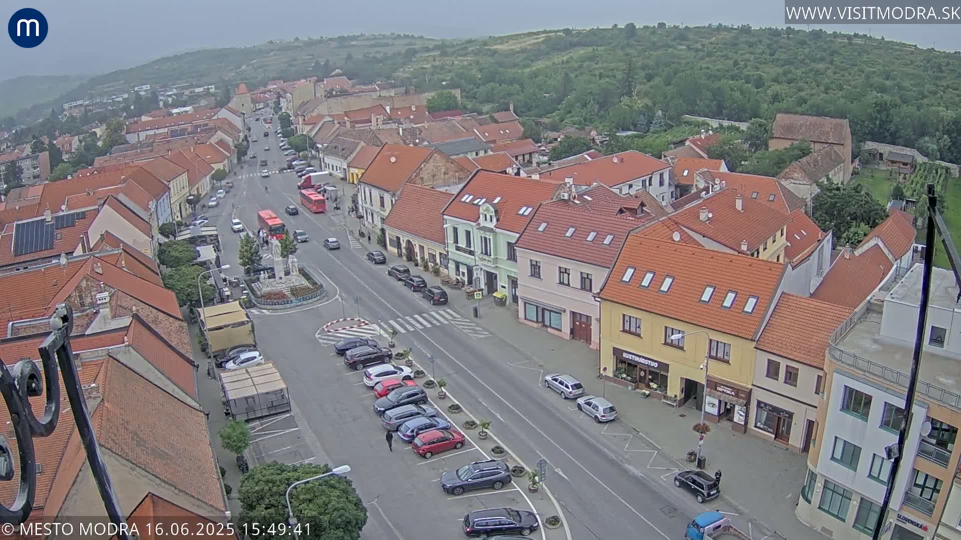 An overcast day reveals a high-angle view of a town square, featuring parked cars, a few pedestrians, and a main road with some traffic, all surrounded by buildings with red-tiled roofs.