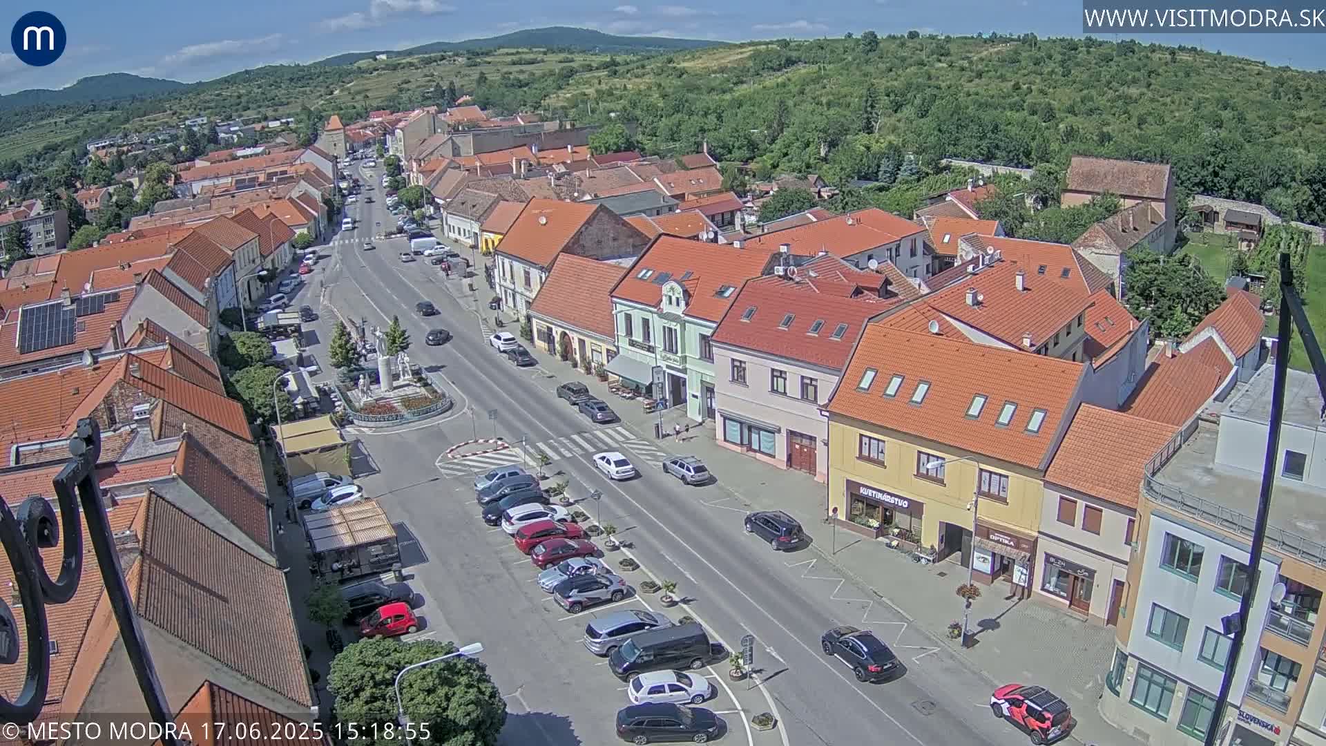 A sunny day reveals a bird's-eye view of a town's main street, lined with parked cars and buildings with red-tiled roofs, all surrounded by lush green hills.