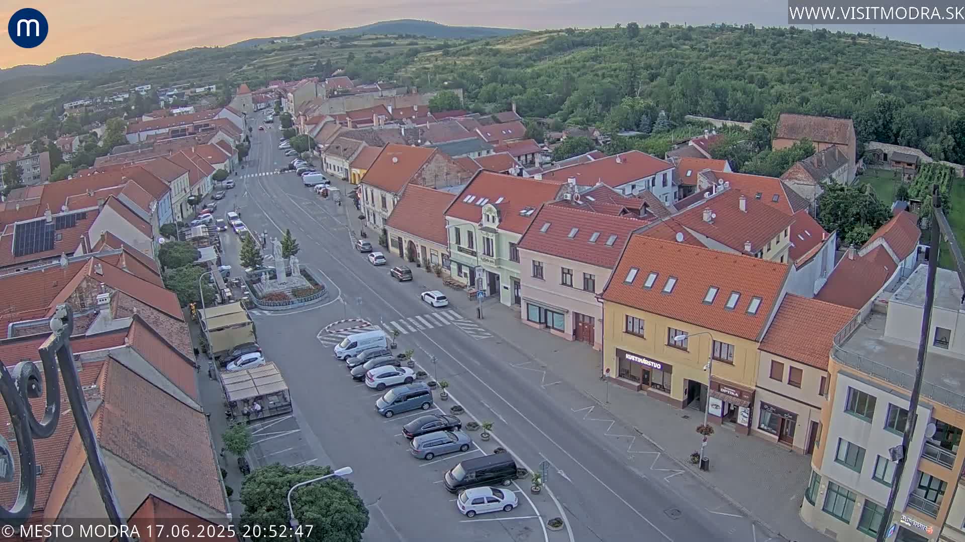 An aerial view shows a town square with parked cars and several buildings under a twilight sky.